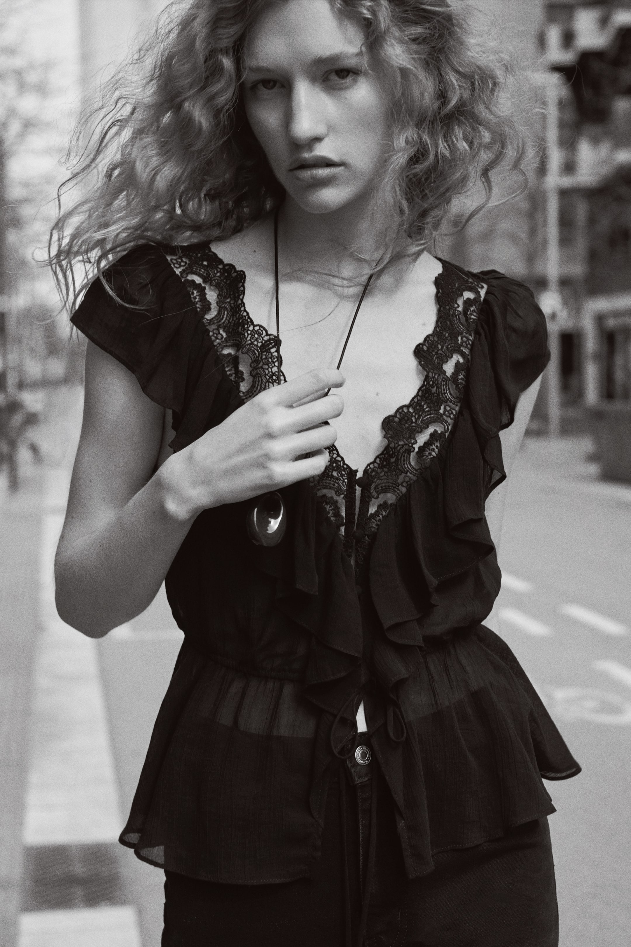 View larger image: A black sheer blouse featuring a deep V-neckline with intricate lace trim and cascading ruffles, along with tie closures at the bust. Designed with short ruffled sleeves and a cinched waist leading to a slightly flared hem.