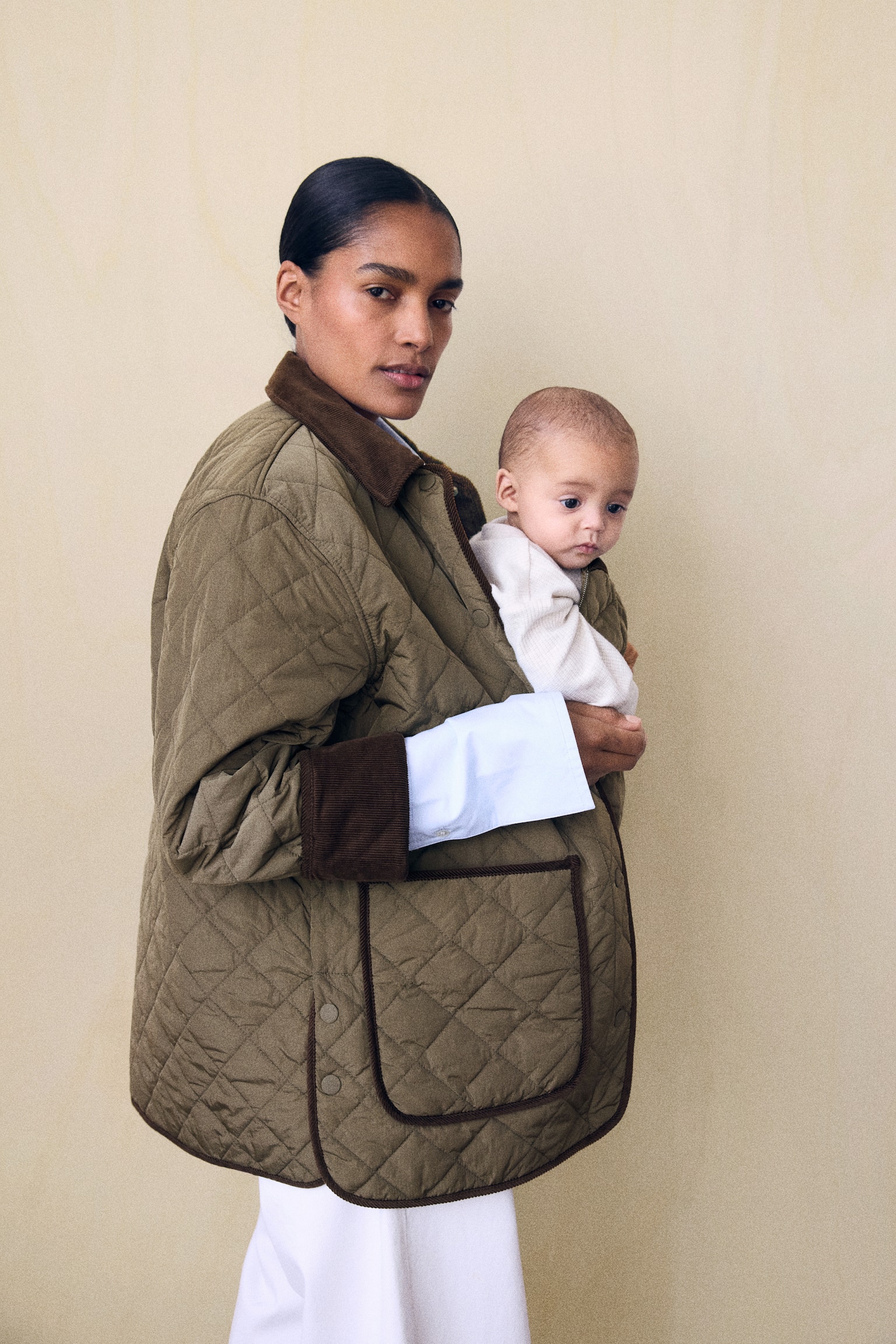 A green quilted jacket with a brown corduroy collar and cuffs is being worn by the model, who is carrying a baby inside the front pouch panel. The model is supporting the baby with one hand, with a white shirt sleeve visible beneath the jacket's cuff. - 1