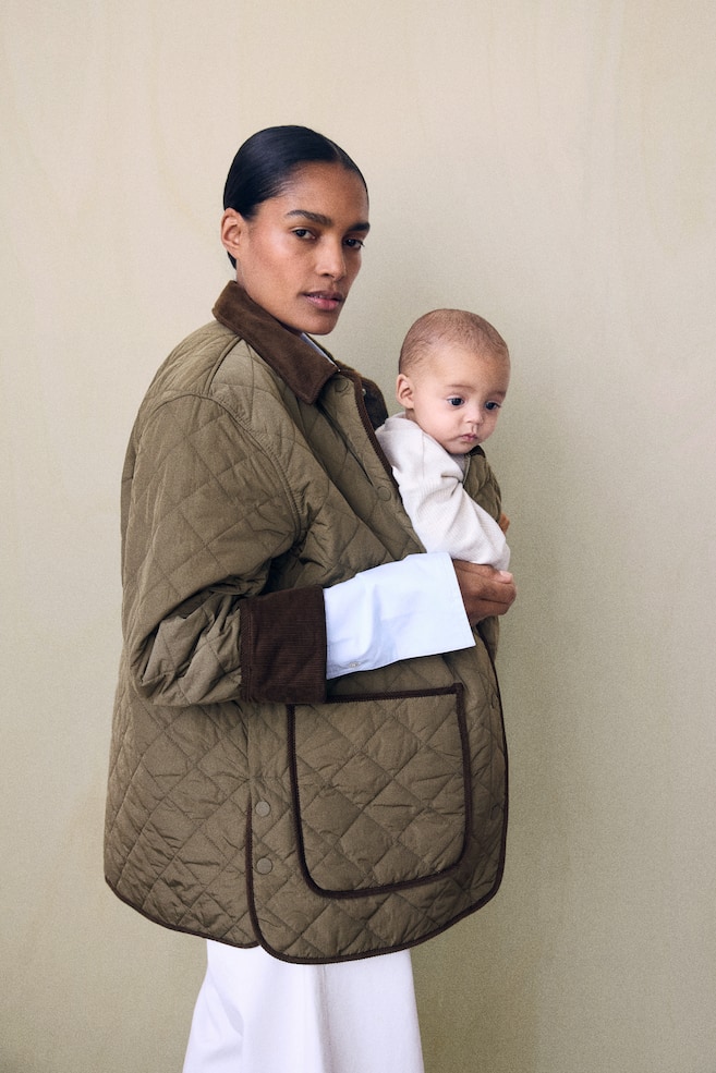 Une veste matelassée vert olive, légèrement rembourrée, est portée par le mannequin, qui porte un bébé à lâintérieur du panneau avant amovible de la veste. La veste présente un col en velours côtelé marron, des poignets retroussés assortis et une grande poche plaquée sur le devant. Le mannequin porte la veste sur une chemise blanche à manches longues et un pantalon blanc. - 1