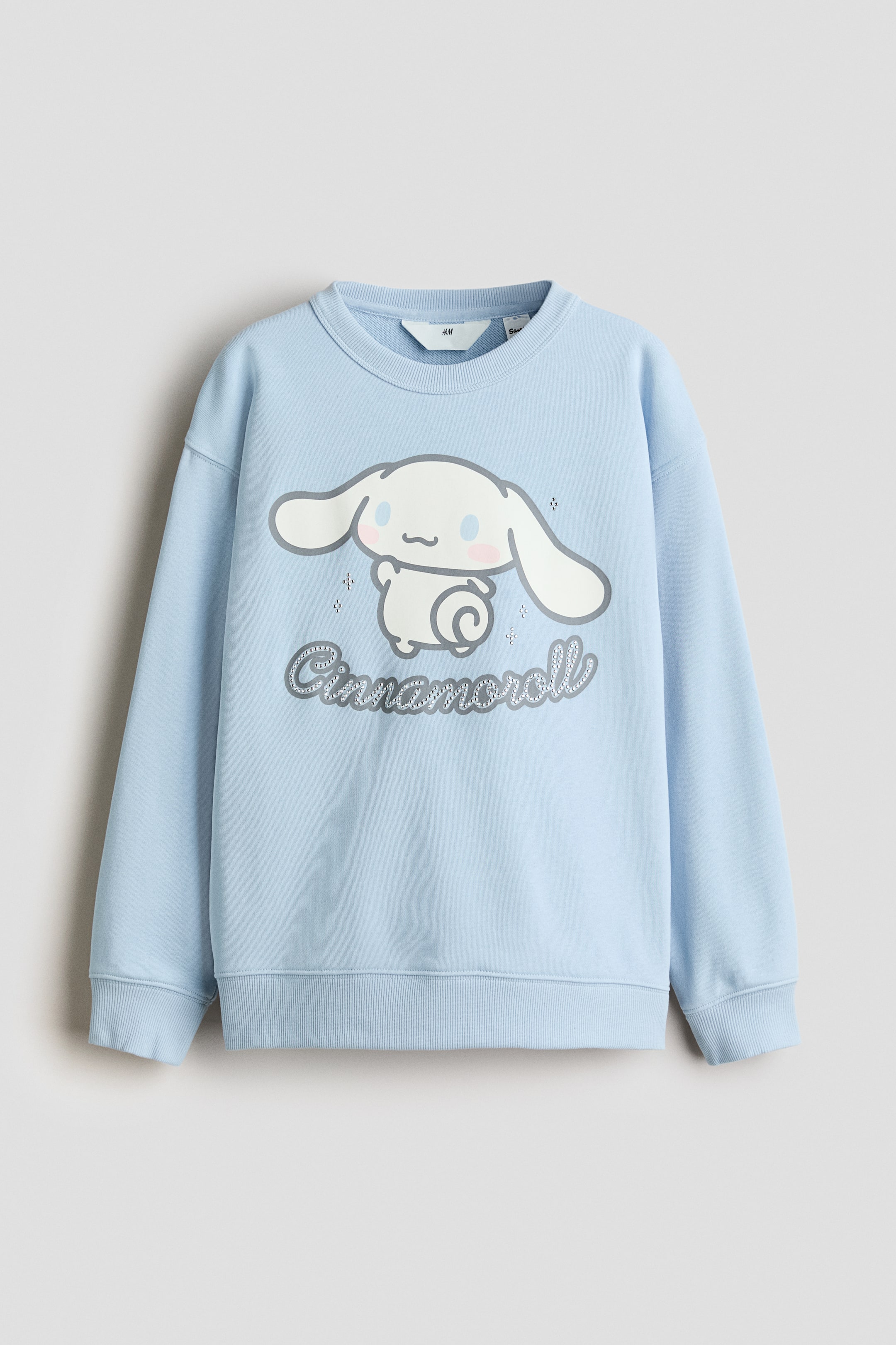 View larger image: A light blue jumper features a large graphic of a white cartoon-style character with big ears, small pink cheeks, and a curly tail, centred on the front, with the word 'Cinnamoroll' appearing in a textured, silvery font below it, accompanied by small sparkling details. It is designed with a ribbed crew neck, ribbed cuffs, and a ribbed hem.