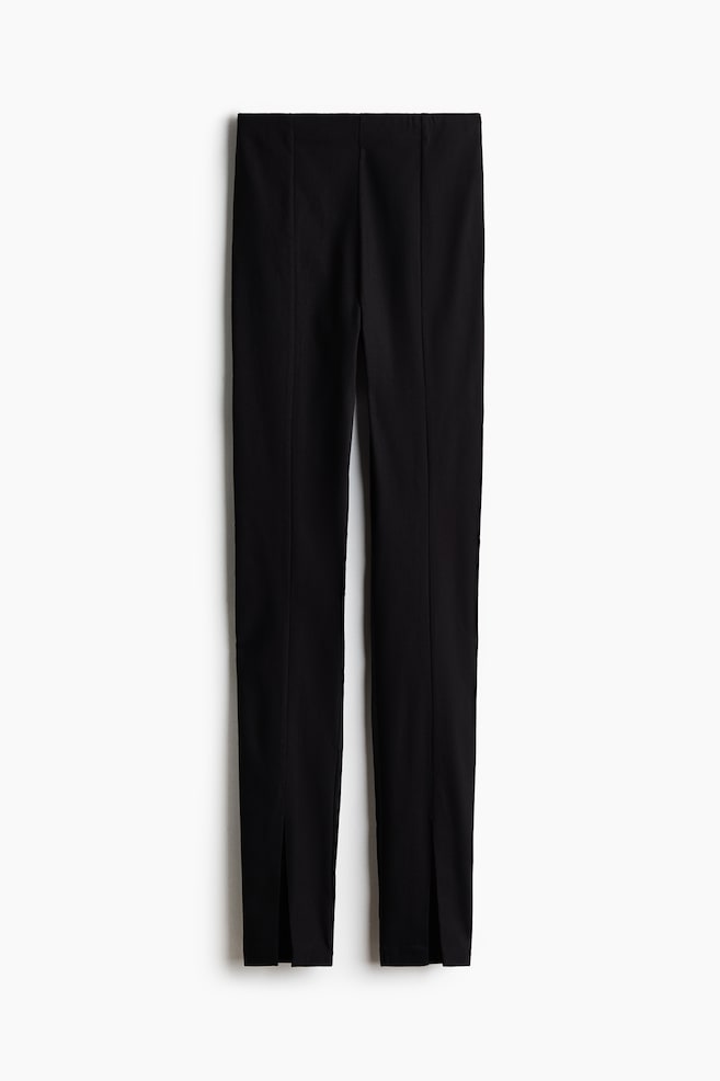 A pair of black slim-fit trousers is shown from the front, featuring a high-rise waistband and defined vertical pintuck seams running down the centre of each leg. The trousers are designed with small split hems at the front of each ankle. - 1