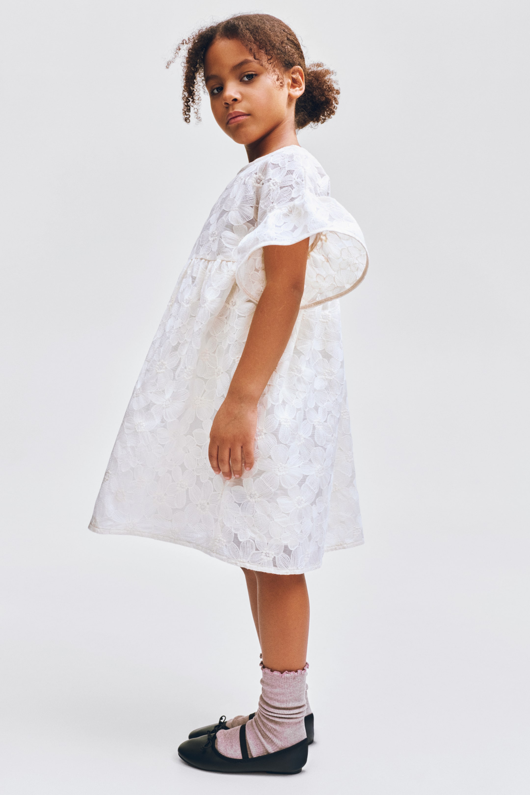 Ampliar la imagen: Un vestido blanco de línea A, visto desde un ángulo ligeramente lateral, presenta un patrón de encaje floral en toda su superficie, mangas cortas y anchas de vuelo, y una costura fruncida en la cintura que crea una falda amplia.