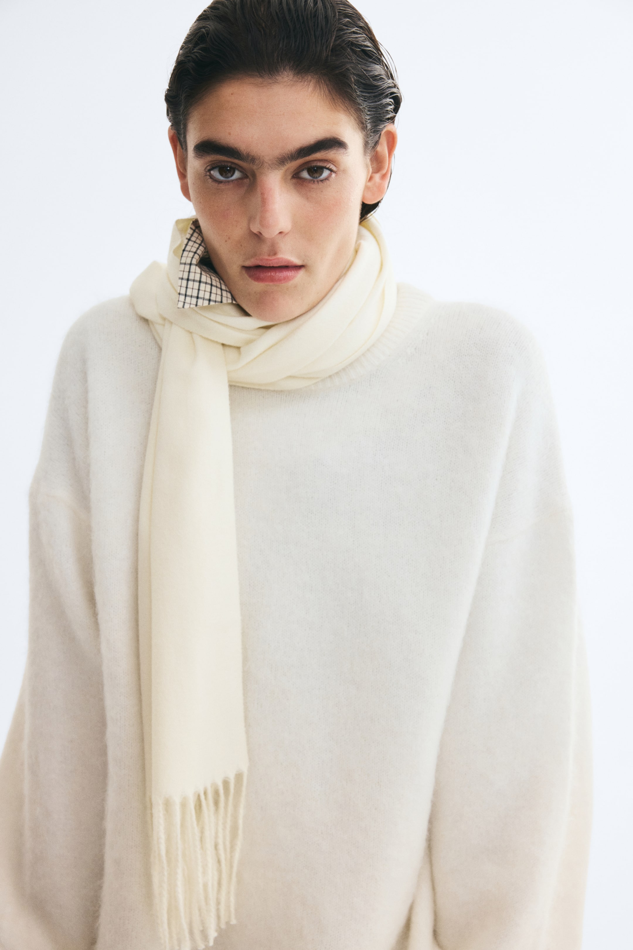 Grösseres Bild ansehen: Ein weich gewebter cremefarbener Schal mit Fransen an den kurzen Seiten wird um den Hals des Models gewickelt getragen. Der Schal ist über einem passenden cremefarbenen Strickpullover gestylt, wobei ein Ende gerade nach vorn herabhängt.