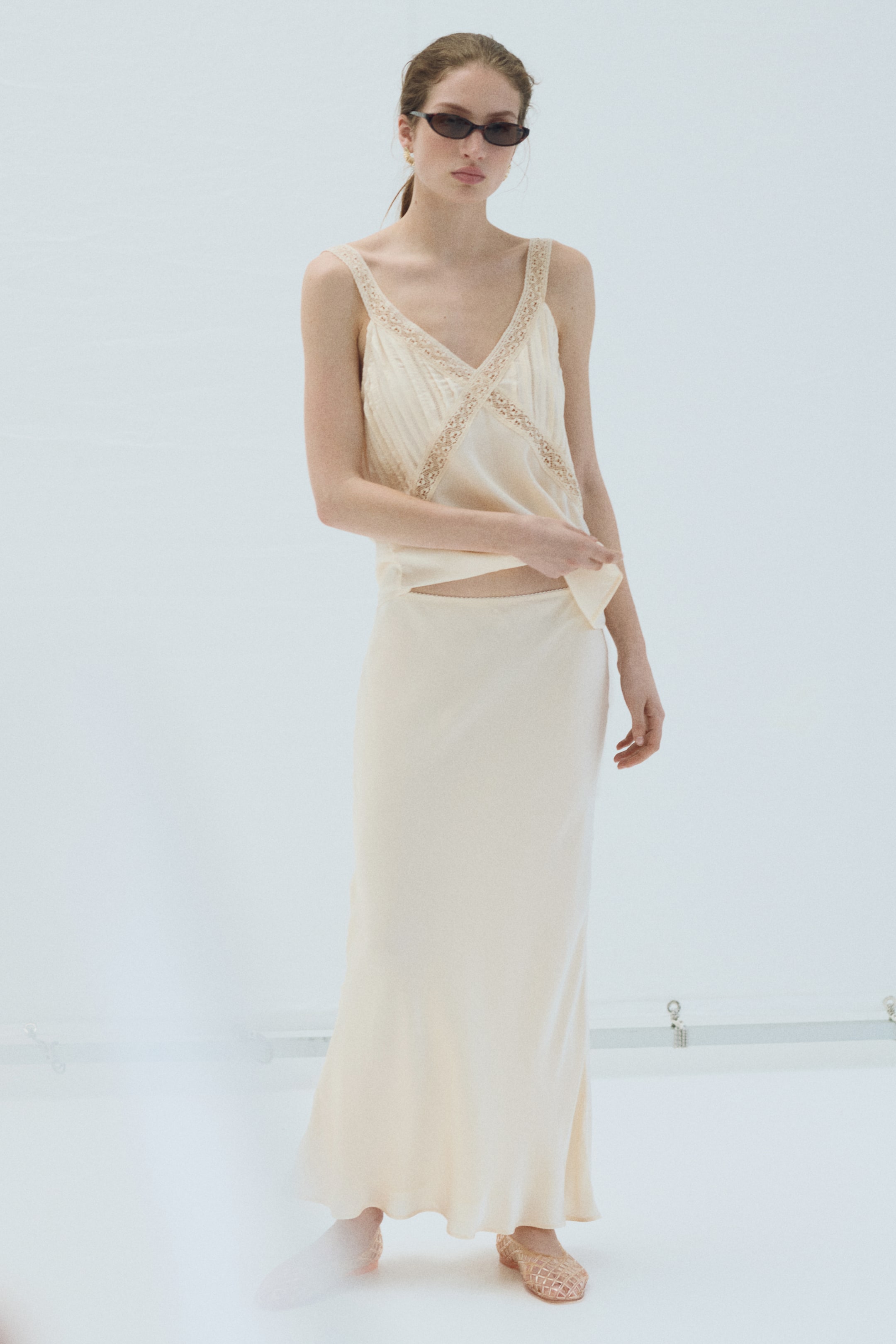 View larger image: The model is wearing a long, fitted slip skirt crafted from a bias-cut viscose and modal weave with a subtle sheen. It boasts a high, elasticated waist adorned with a visible picot trim and gently flares at the hem. The model lifts the top to showcase the waistband.