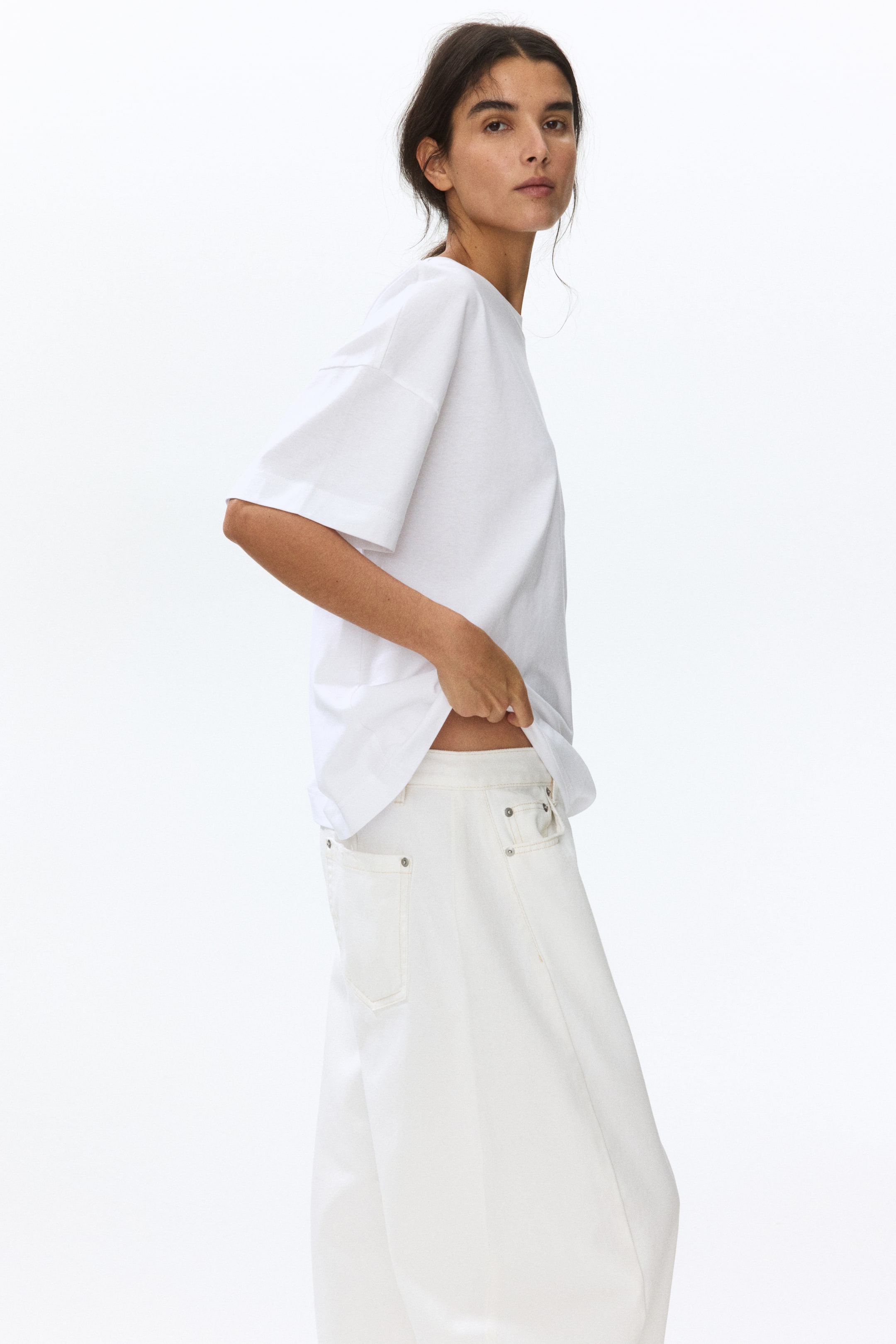 View larger image: A white relaxed-fit t-shirt is showcased with wide short sleeves and a crew neck, presented from a slight side angle. The outfit is paired with white wide-leg denim pants, featuring back pockets adorned with a decorative ring and a high waist. The t-shirt is slightly lifted to reveal the waistband of the pants, offering a stylish and casual look.