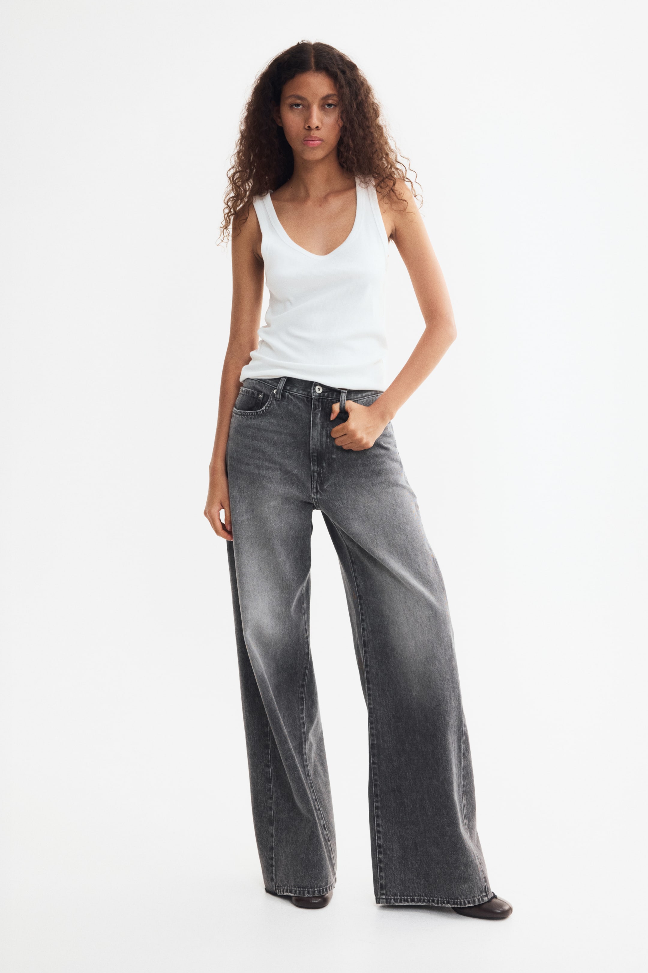 Grotere afbeelding bekijken: De model draagt een vervaagde zwarte denim jeans met wijde pijpen en een normale taille, terwijl ze staat met één hand rustend bij de voorzak. De jeans heeft een rechte, losse pasvorm en wordt gecombineerd met een witte tanktop.