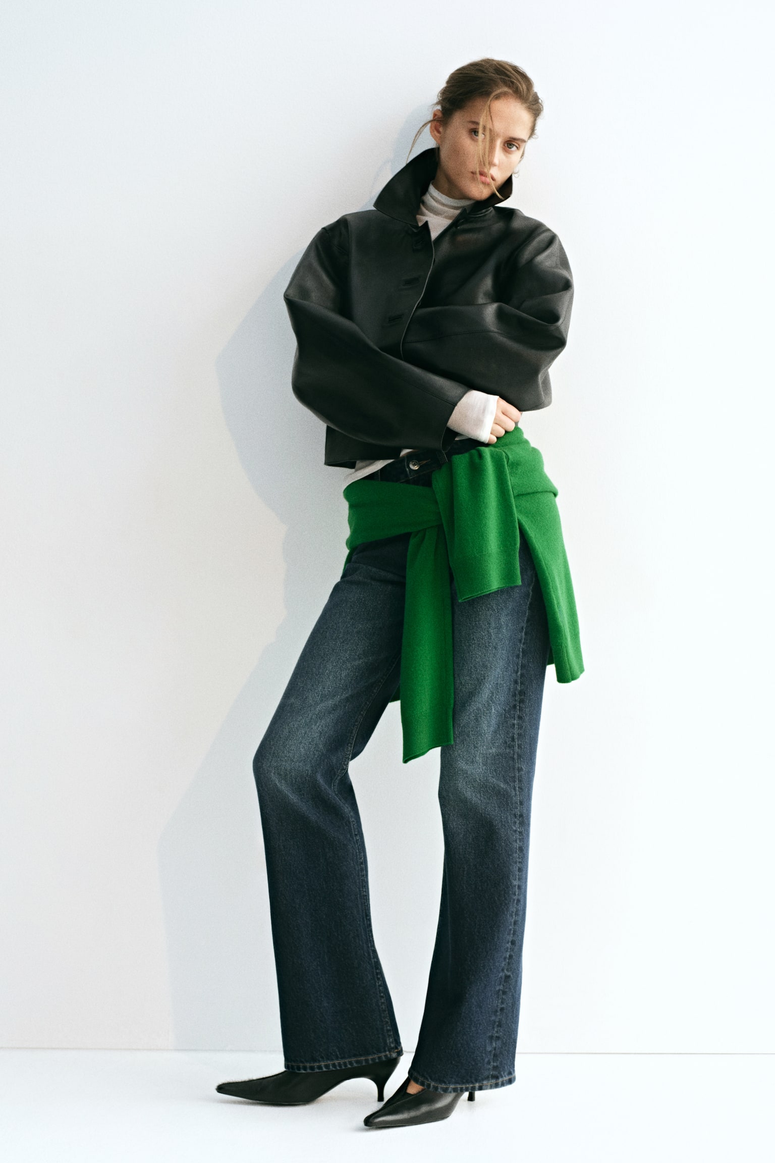 La modella indossa jeans in denim blu scuro, con vita alta, vestibilitÃ  slim sulle cosce e fondo bootcut svasato. I jeans sono abbinati a un maglione verde brillante annodato in vita, una giacca in pelle nera e dÃ©colletÃ© nere a punta. - 1