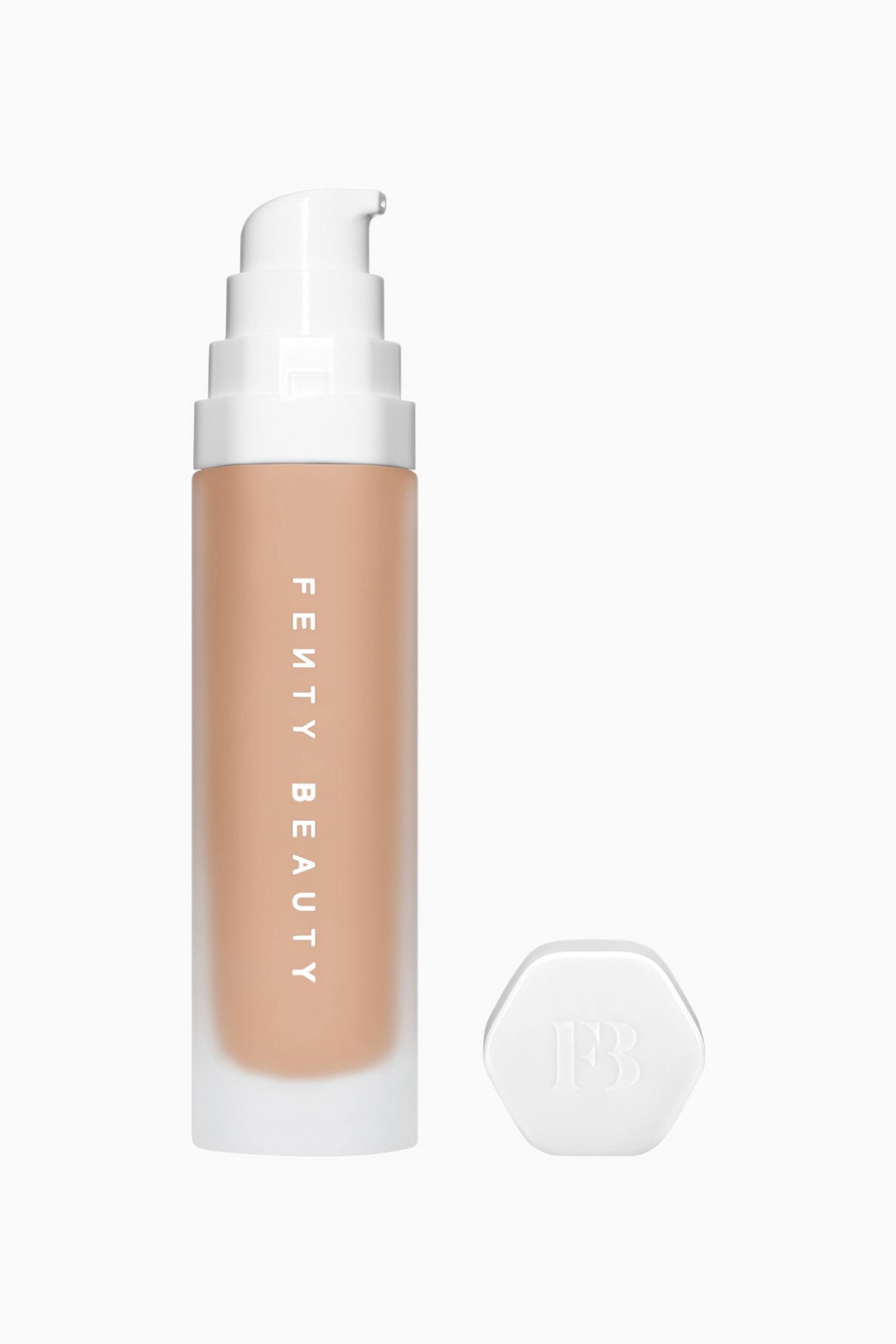 Visa större bild: Soft'lit Naturally Luminous Longwear Foundation står upprätt och visar upp en frostad cylindrisk flaska fylld med en medium-tan-färgad vätska, med den vertikala logotypen "FENTY BEAUTY" i vitt. En vit, trappstegsformad pumputdelning pryder flaskans topp, medan en separat vit sexkantig kork med präglad logotyp ligger bredvid.