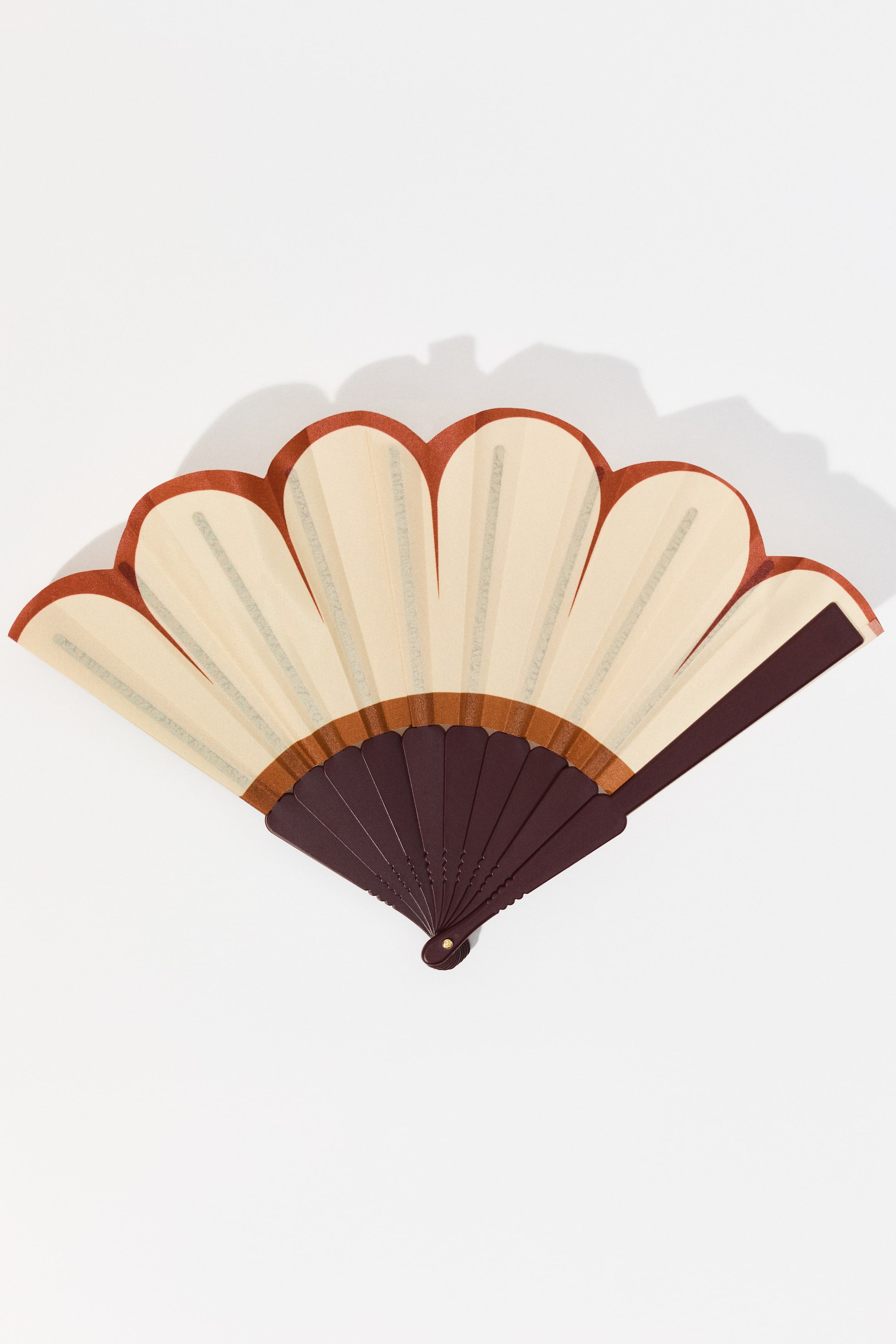 הצגת תמונה גדולה יותר: מניפה מתקפלת מוצגת כשהיא פתוחה חלקית, נפרשת מהפינה הימנית התחתונה. היא מציגה עלה בצבע קרם עם קצוות מסולסלים בצבע חום-אדמדם, וקווים אנכיים דקים בצבע אפור בהיר נראים על כל מקטע. מקלות שמירה כהים וריבועים פנימיים בצבע חום כהה יוצרים את מבנה המניפה.