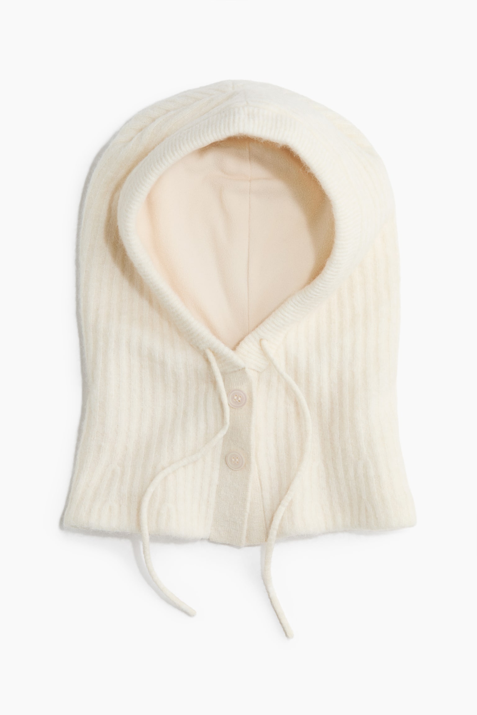 Une capuche tricotÃ©e de couleur crÃ¨me est prÃ©sentÃ©e de face, montrant une texture extÃ©rieure cÃ´telÃ©e et un intÃ©rieur doublÃ© de tissu lisse. Elle comporte une fermeture Ã  boutons sur le devant avec deux boutons au niveau du cou, ainsi que des cordons de serrage assortis qui pendent de lâouverture de la capuche.