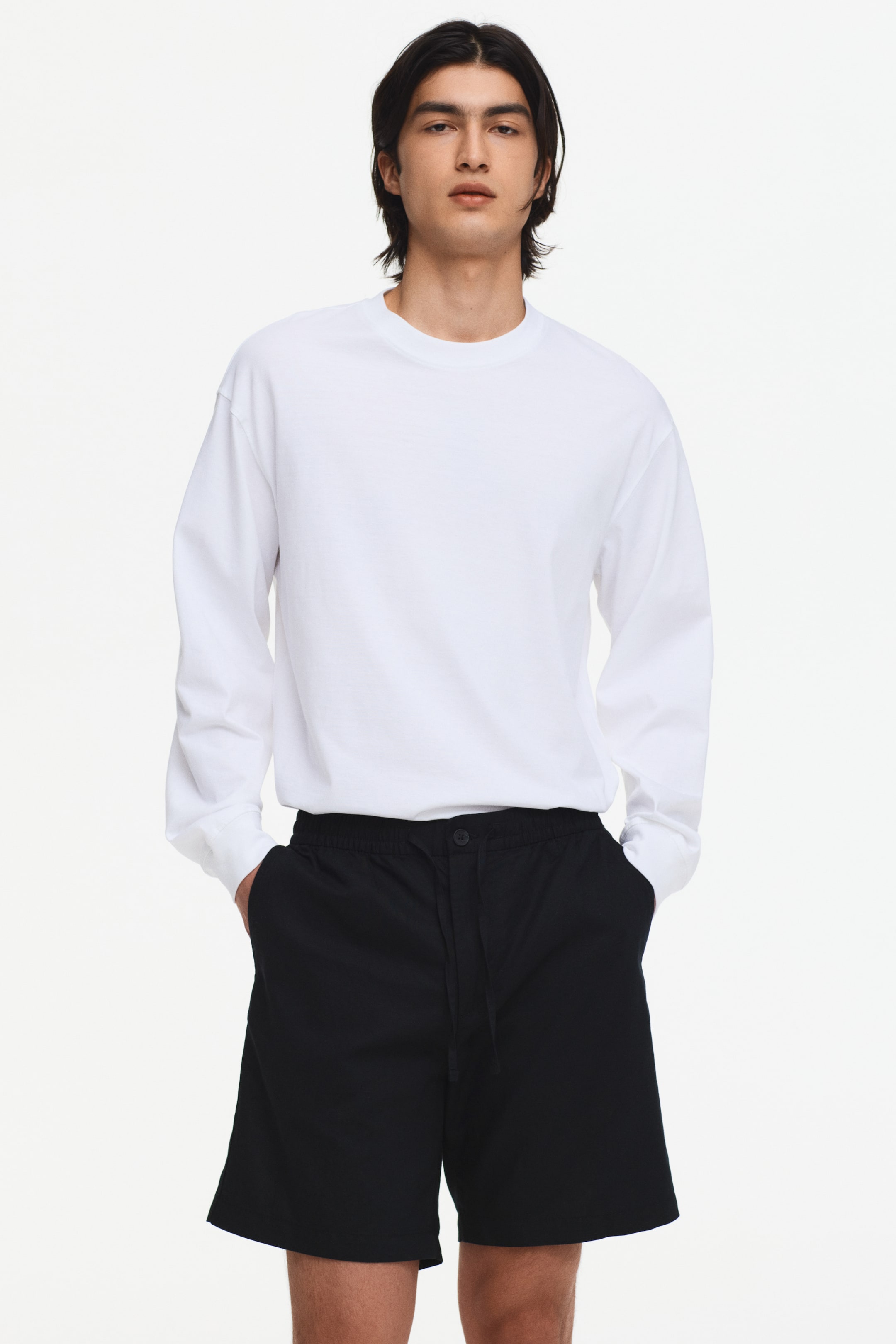 View larger image: A white long-sleeved tee showcases a crew neck and a relaxed, slightly oversized fit with long sleeves. Black shorts exhibit an elasticated waist with a drawstring and button closure, alongside side pockets where hands rest inside, all captured from a frontal perspective.