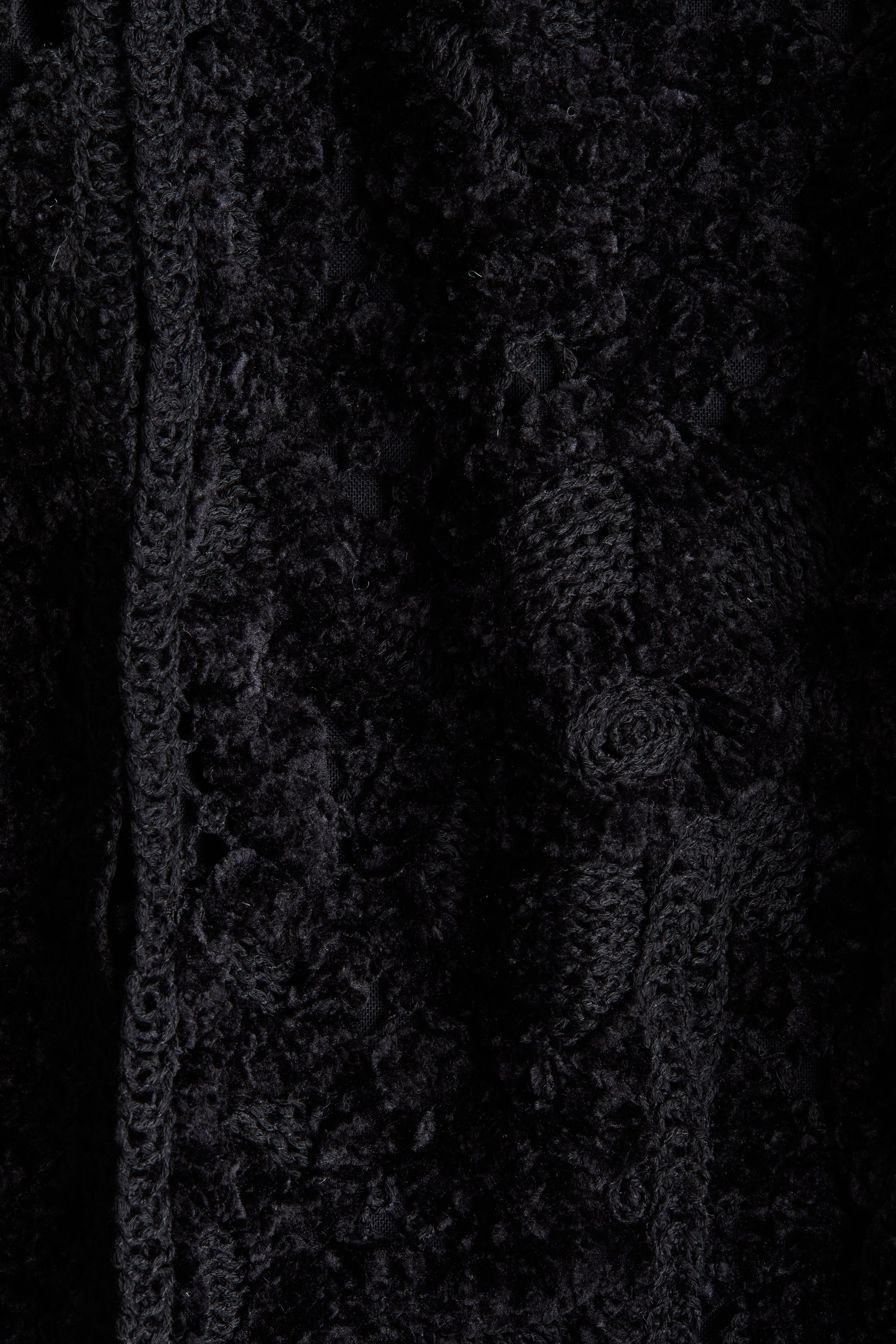 Größeres Bild ansehen: Ein schwarzer Stoff weist eine dichte, lockige, geschlungene Textur auf. Ein vertikaler Streifen mit offenen, ineinandergreifenden Details verläuft entlang der linken Seite, und innerhalb der Haupttextur erscheinen deutliche rosettenartige Muster.
