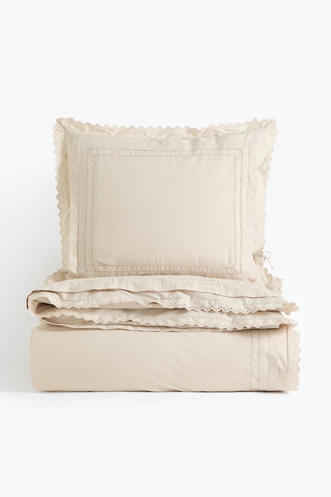 Um conjunto de roupa de cama bege apresenta uma fronha decorativa virada para a frente, empilhada sobre uma capa de edredão dobrada. Ambos os artigos apresentam um acabamento em renda com bico ao longo das bordas e delicados detalhes em pesponto decorativo. - 1