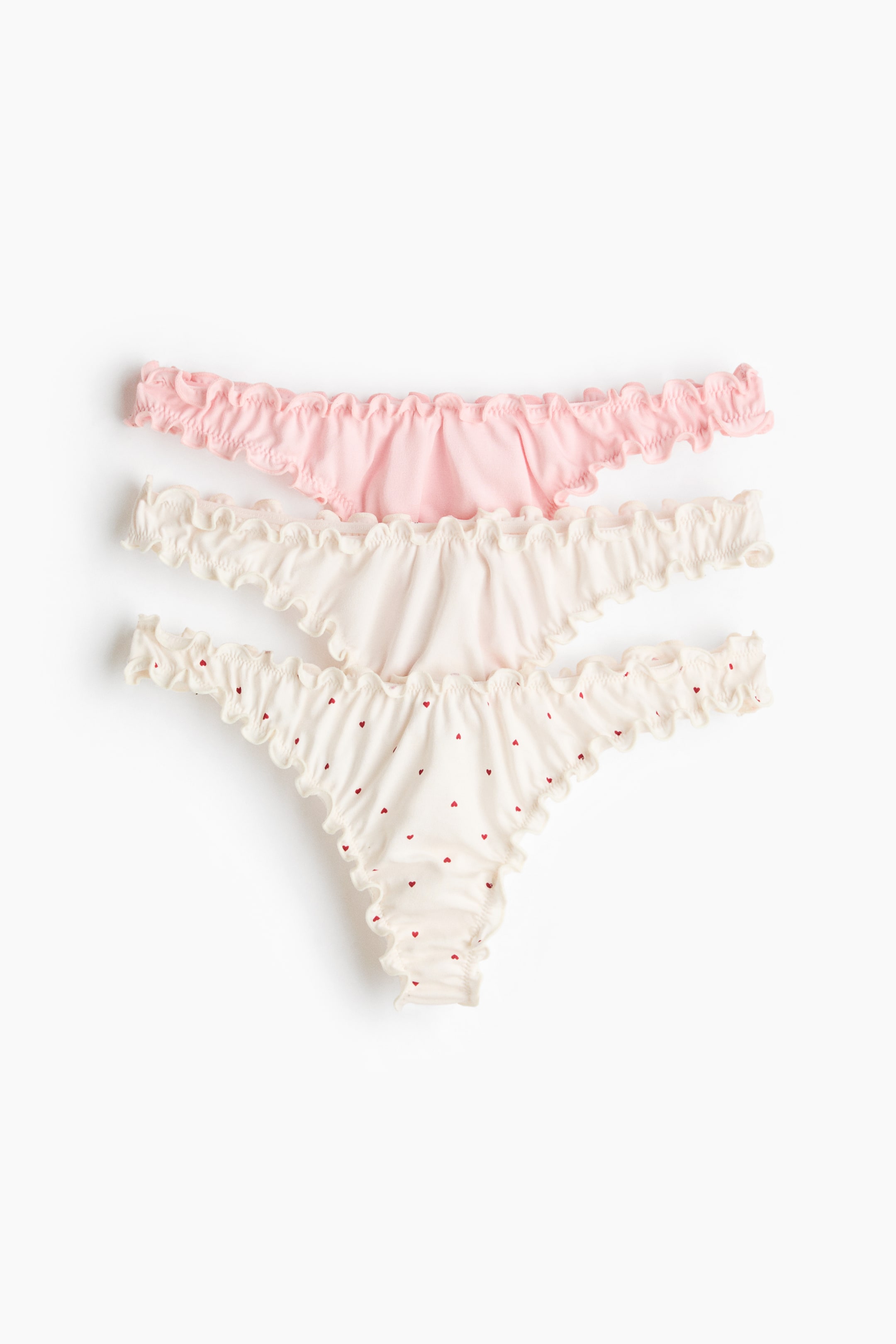 Vis større bilde: Tre stringtruser ligger flatt og frontvendt, med volangkantede, overlockede kanter langs midjen og benåpningene, samt en lav, V-formet midje. De vises i rosa, off-white og off-white med små røde hjerte-mønstre.