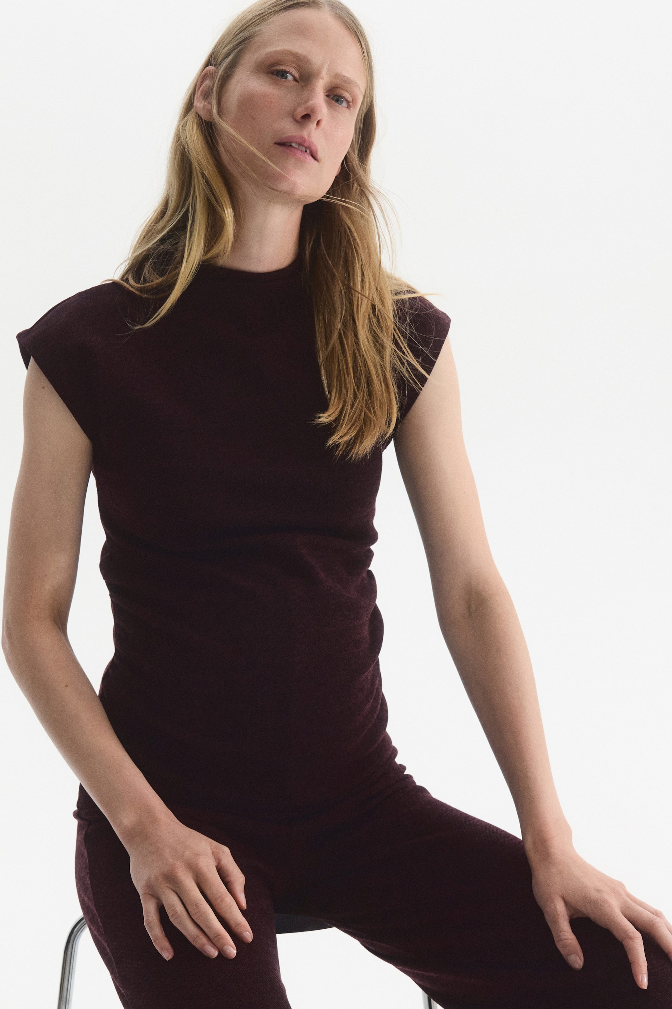 View larger image: A dark burgundy jersey set, featuring a high-neck top with cap sleeves and matching pants, is worn by the model. The model is seated on a stool while wearing the set.