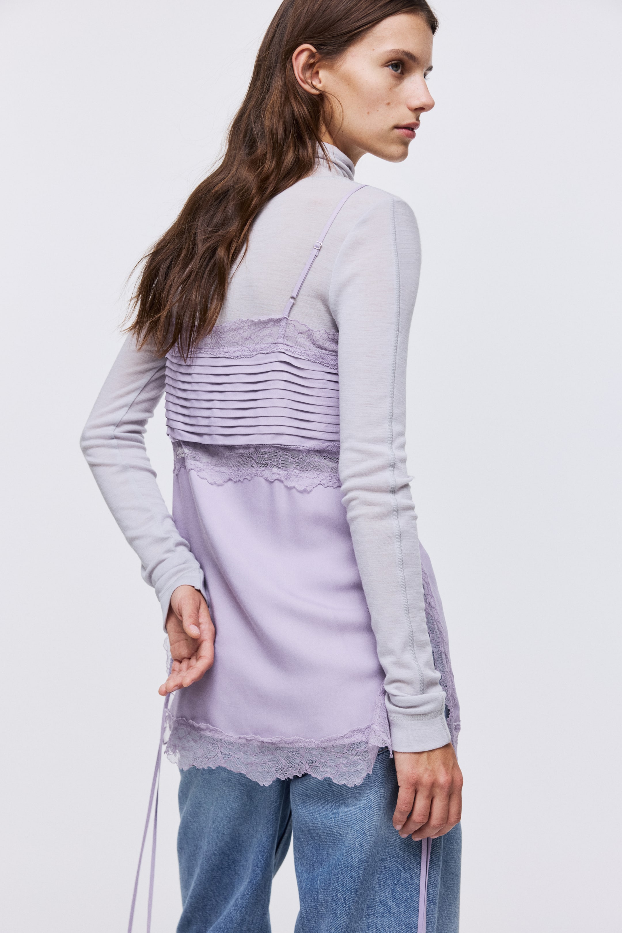 View larger image: A lilac viscose long top, featuring lace edges and insets, a pleated section over the bust, and adjustable spaghetti shoulder straps, is being worn by the model. The top is layered over a light gray long-sleeved turtleneck, and its high side slits have narrow ties that the model's hand is touching.