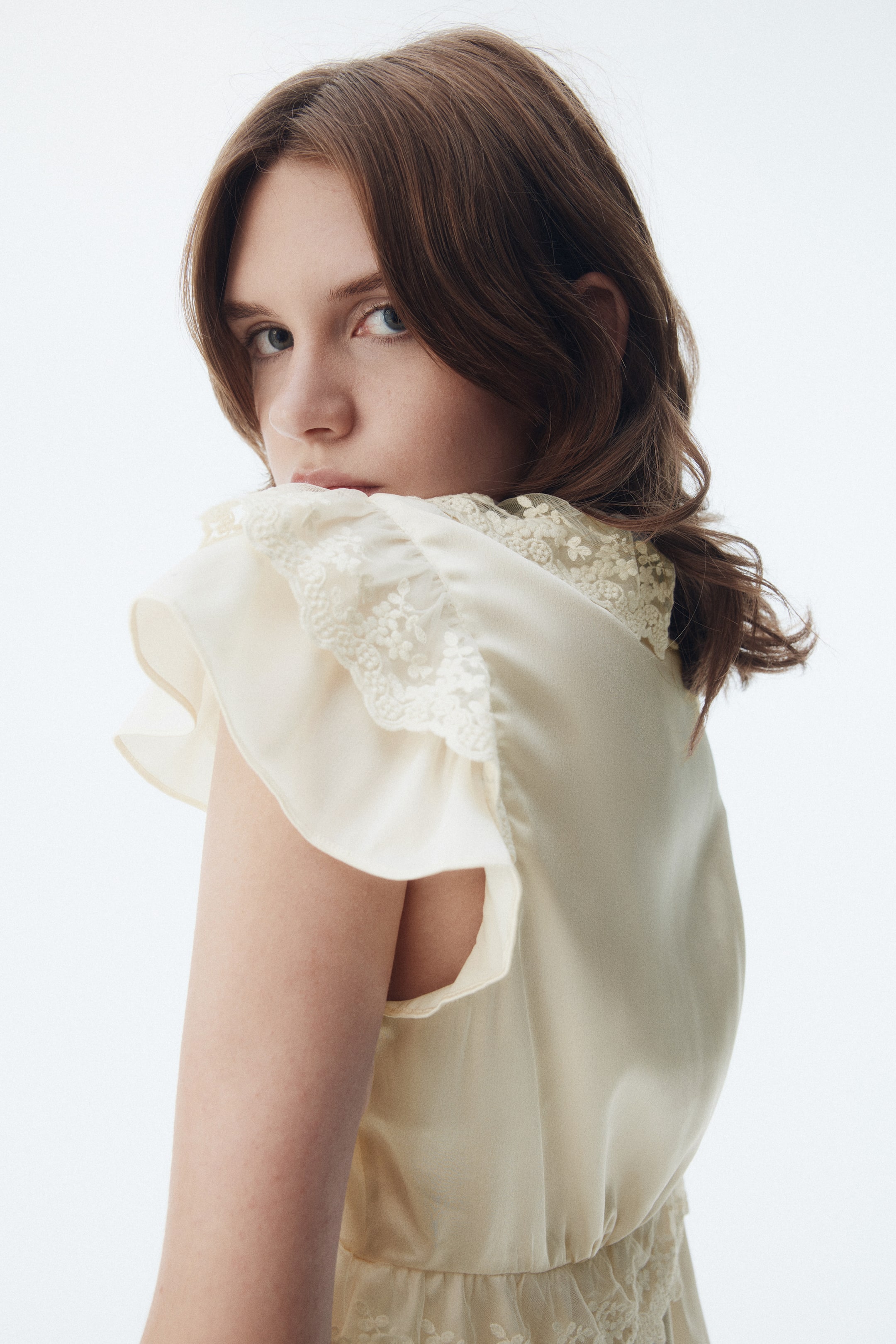 View larger image: A cream-colored dress, seen from a back-side angle, is featuring ruffled cap sleeves with delicate lace trim. The dress is showcasing intricate lace details around the neckline and a gathered waist, with an additional lace panel visible below.