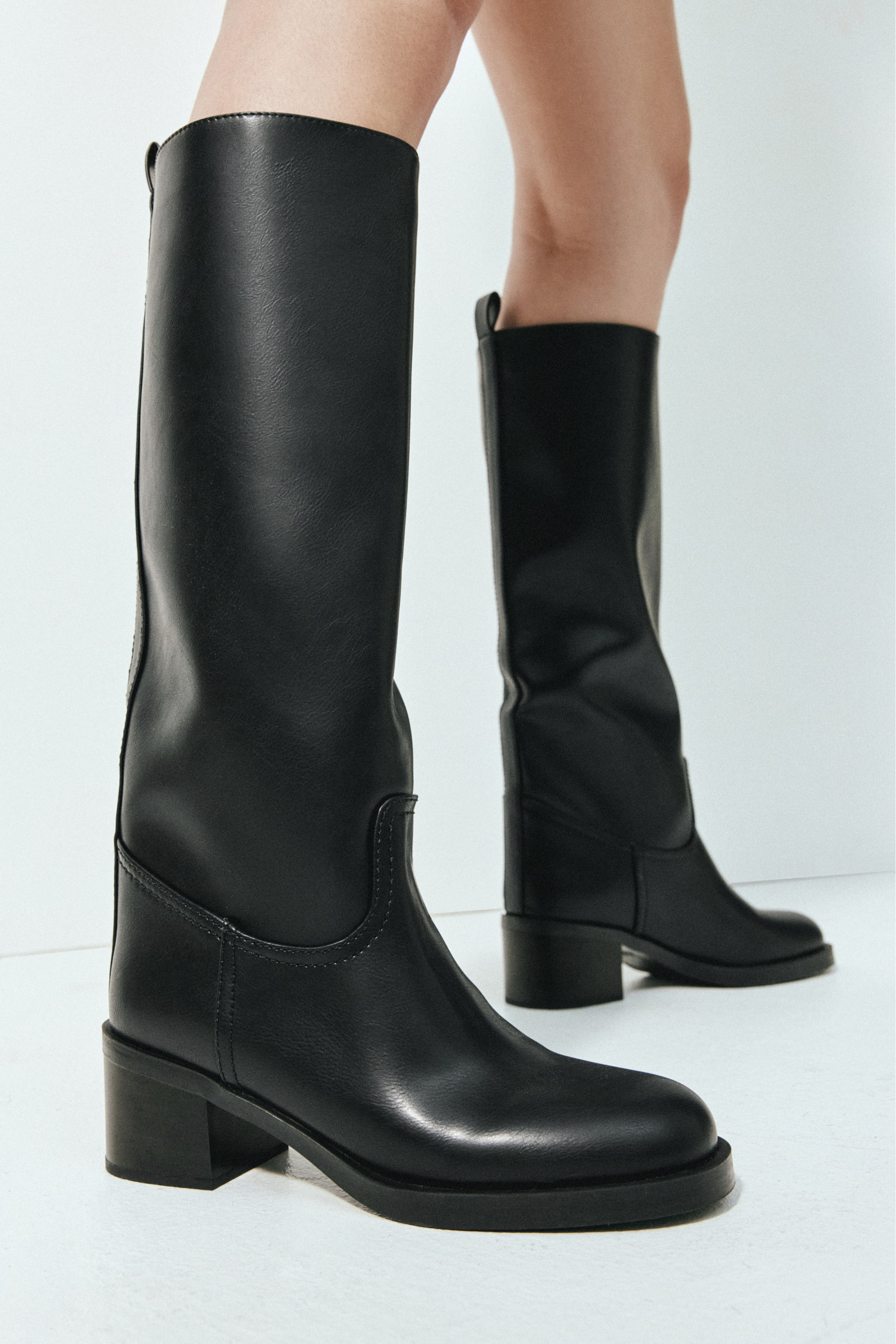 Größeres Bild ansehen: Schwarze, kniehohe Lederstiefel mit abgerundeter Zehenpartie, niedrigem Blockabsatz und Zuglaschen an der Rückseite der Schäfte werden von dem Model getragen. Die Stiefel haben eine strukturierte Silhouette mit dezenten Nahtdetails am Knöchel und Fuß.