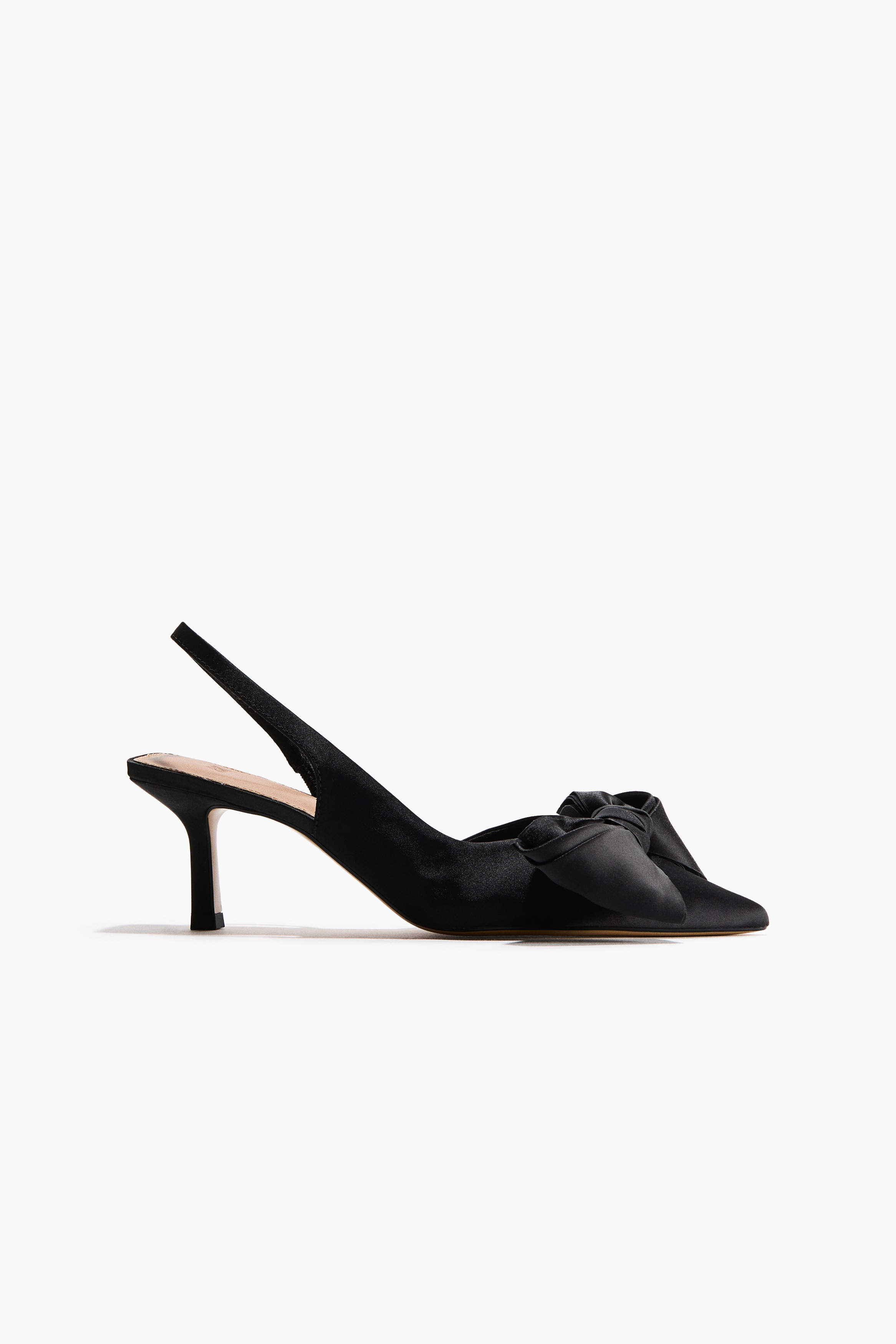A black satin shoe featuring a pointed toe and a large, decorative bow at the front is being worn by the model. The shoe is styled with a draped black garment visible around the leg.