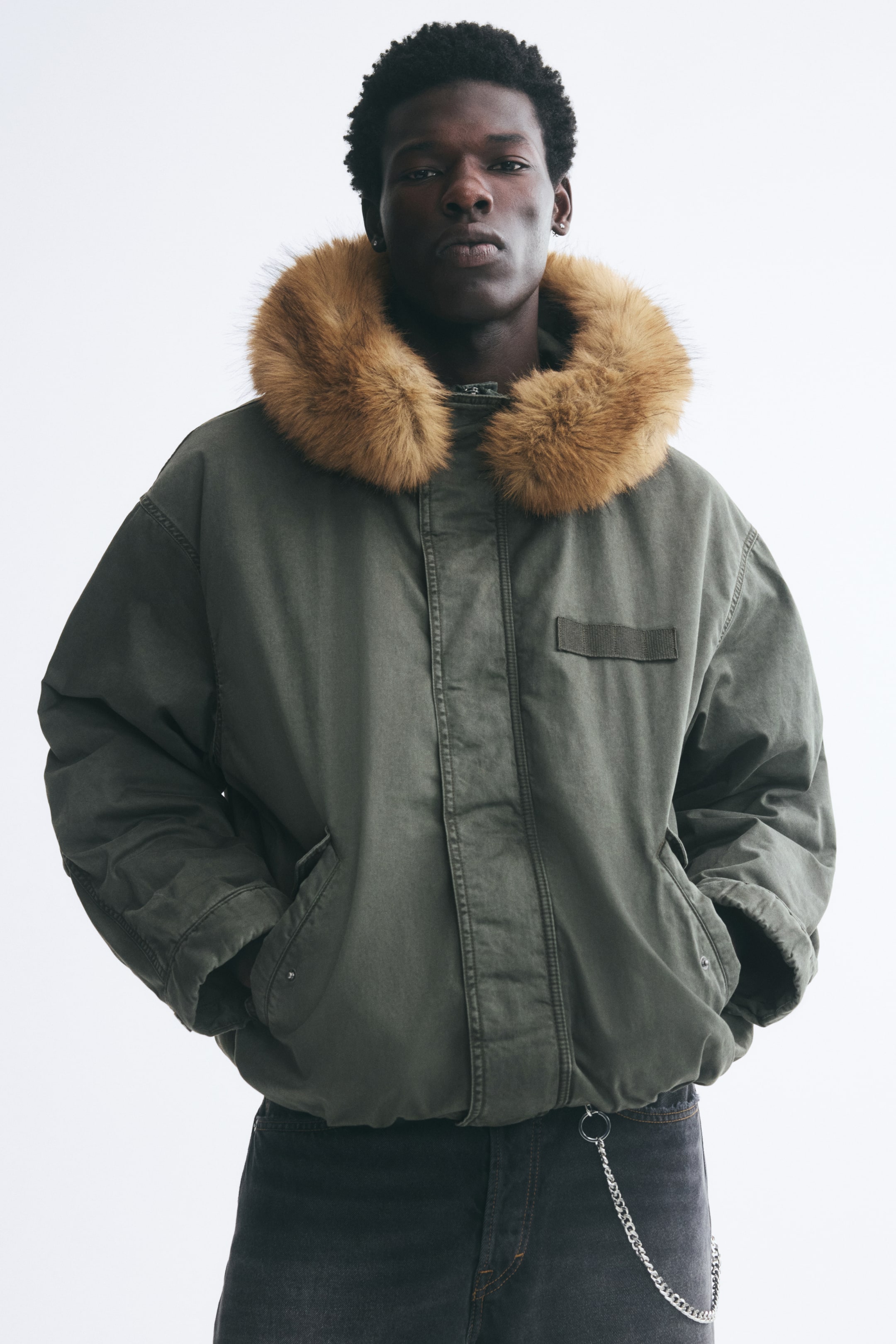 Visa större bild: En olivgrön vadderad parka med en fluffig, ljusbrun fuskpälskrage bärs av modellen. Modellen står med händerna i de främre fickorna på den uppdragna jackan, som kombineras med mörkgrå jeans.