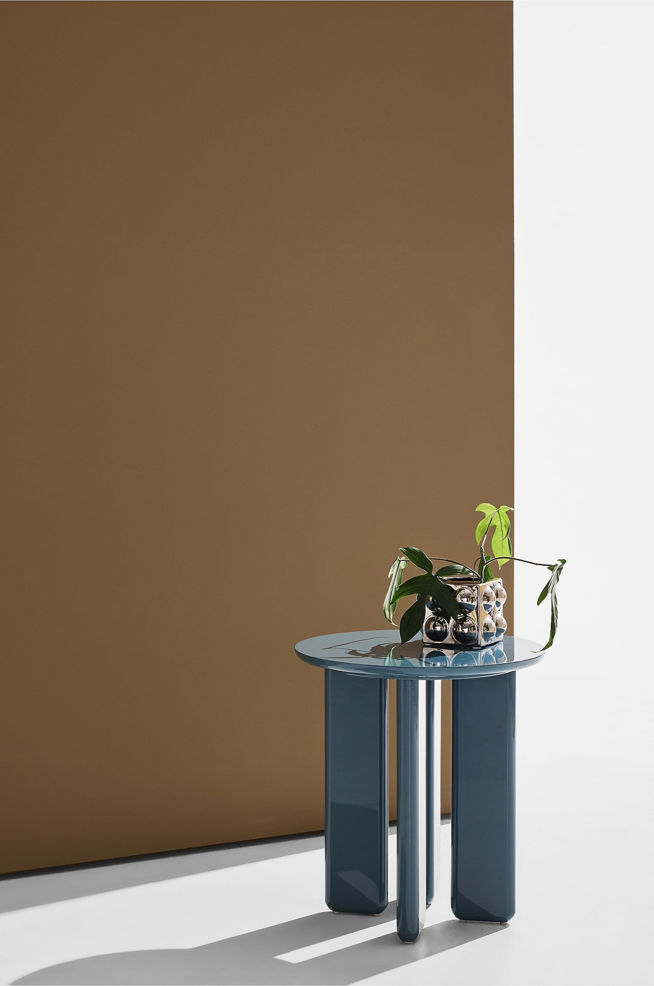 Visa större bild: Ett runt, mörkt teal sidobord är placerat i en lätt vinkel, vilket framhäver dess glansiga yta och tre tjocka, rundade ben. En växt i en metallisk, bubbeltexturerad kruka vilar på bordsskivan.