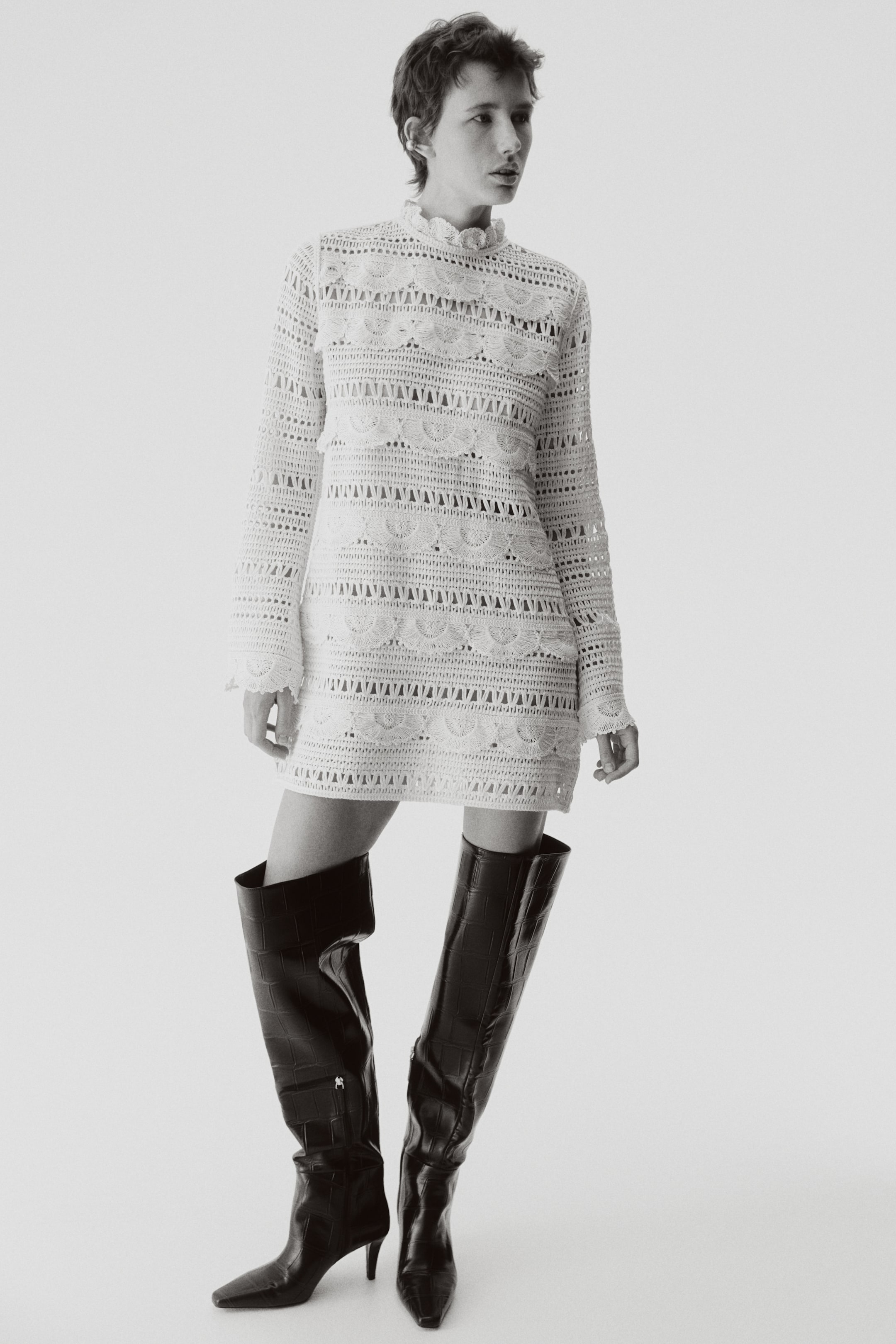 View larger image: A white crochet mini dress is presented from a front-facing angle, featuring a high ruffled neckline and long sleeves. It showcases intricate horizontal bands of openwork patterns, including scalloped motifs and geometric designs, throughout the body and sleeves.