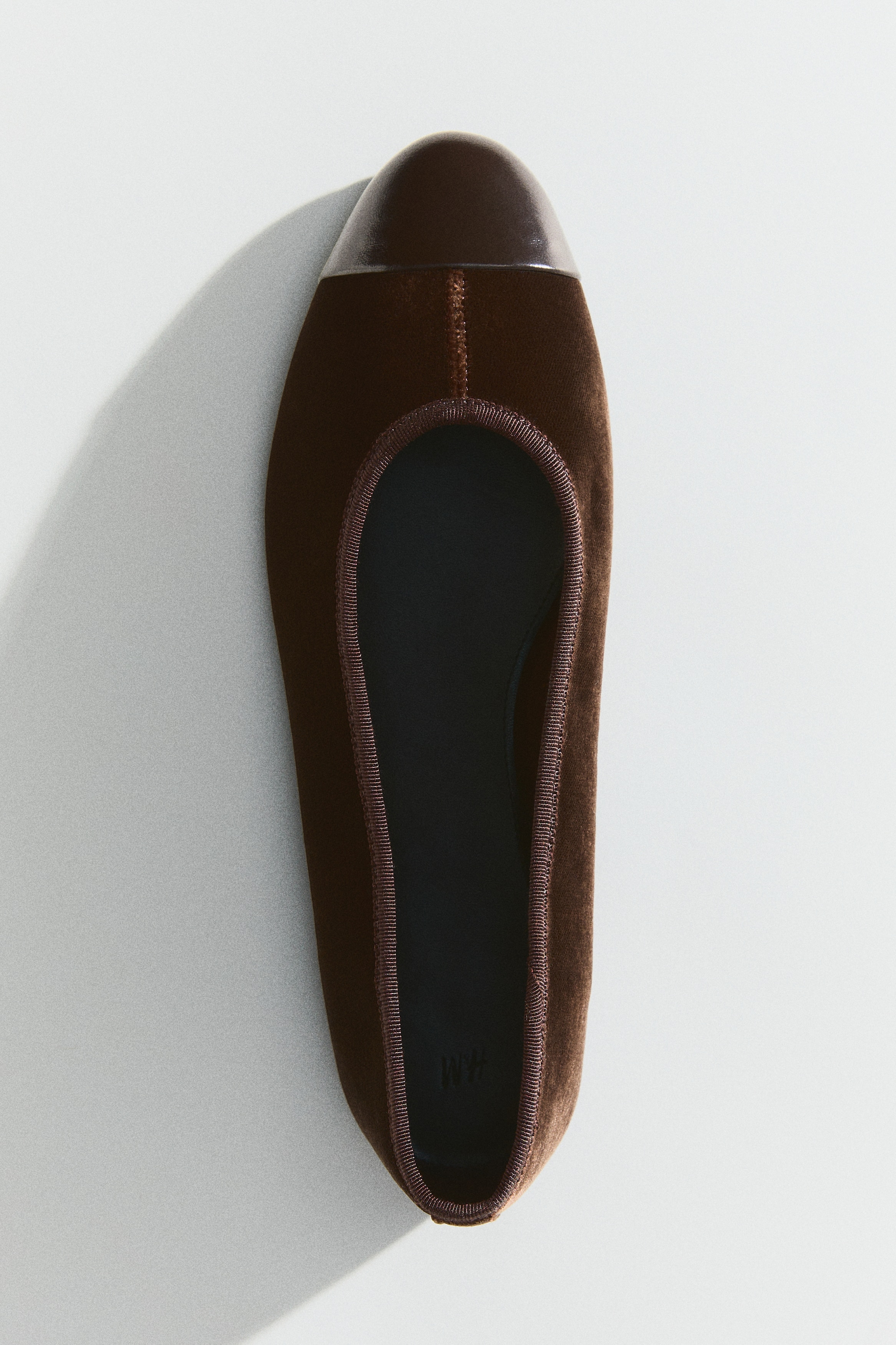 Ein Paar braune Ballerinas wird präsentiert, wobei jeder Schuh samtige Obermaterialien und eine abgerundete, dunkelbraune Lacklederspitze aufweist. Ein dünner, gerippter brauner Besatz umrahmt die Öffnung, und eine schwarze Innensohle liegt innen.