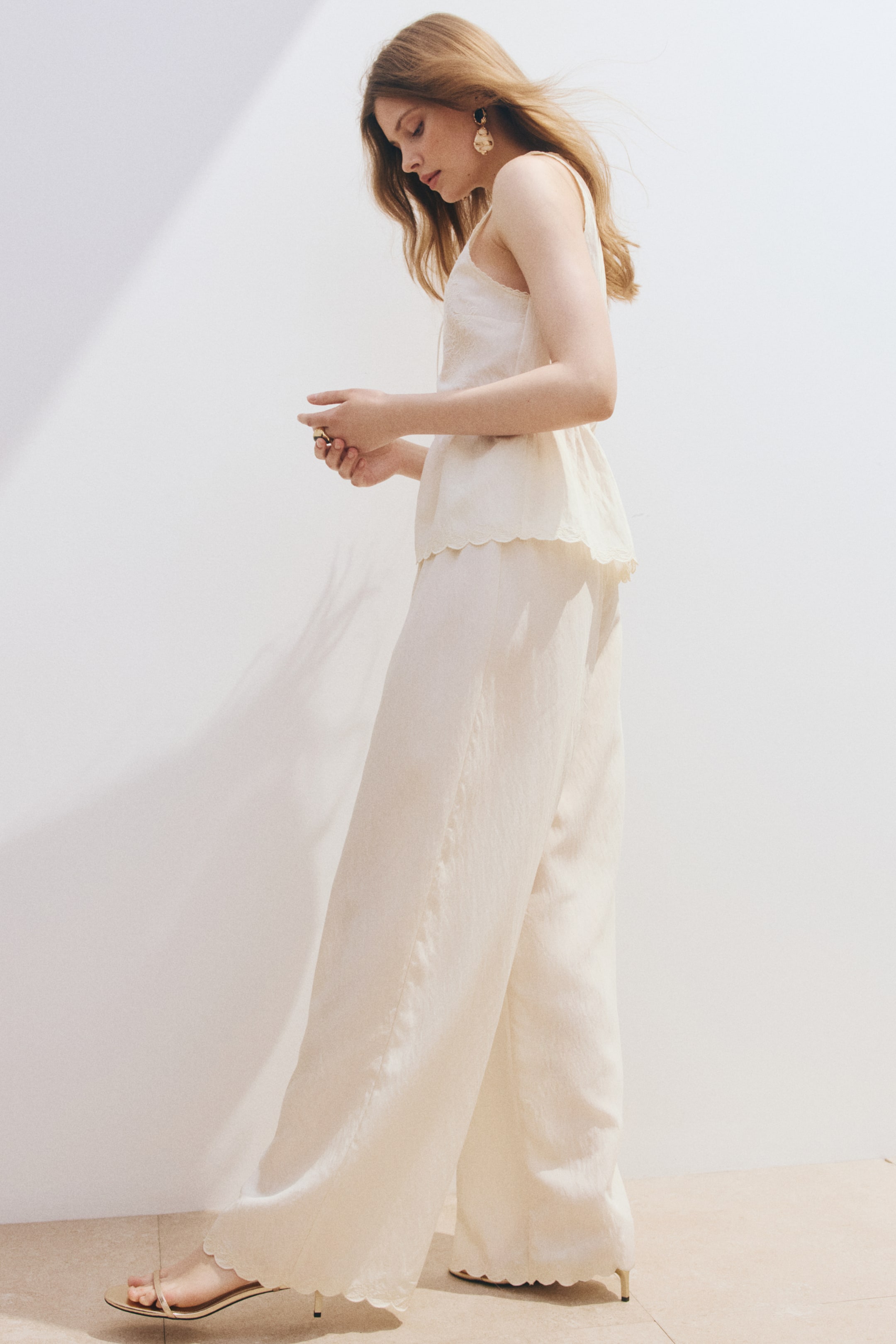 View larger image: A cream-coloured matching set is showcased from a side angle. The sleeveless top boasts thin straps and a scalloped peplum hem, while the wide-leg trousers display a subtle crinkled texture and a matching scalloped hem.