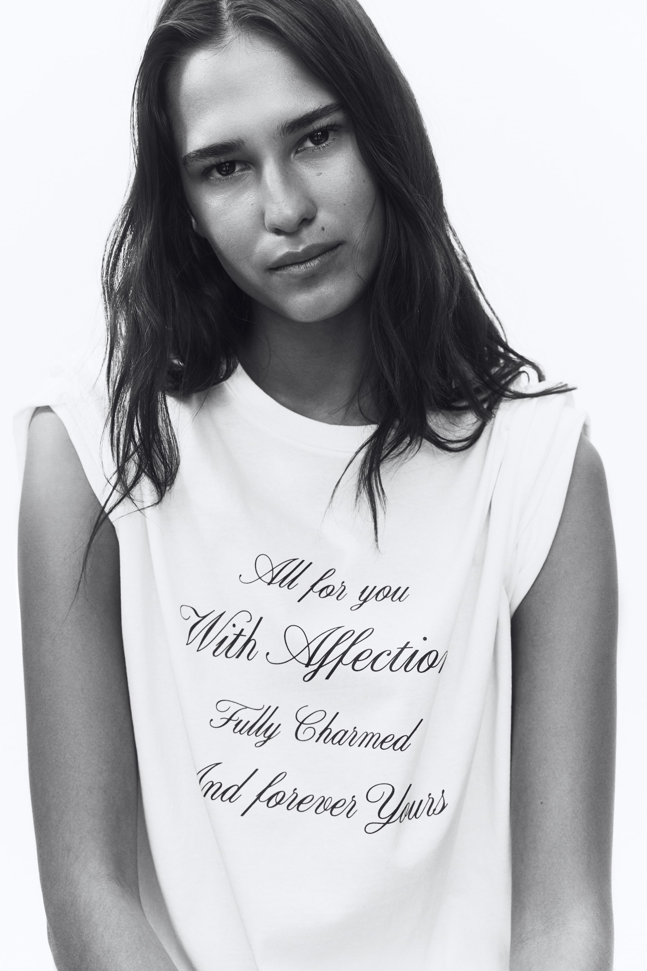 拡大画像を表示: モデルが着用しているのは、コットンジャージー素材のオーバーサイズTシャツです。リブ付きのネックライン、ドロップショルダーが特徴で、フロントには「All for you With Affection Fully Charmed And forever Yours」と書かれた黒いスクリプト文字がプリントされています。