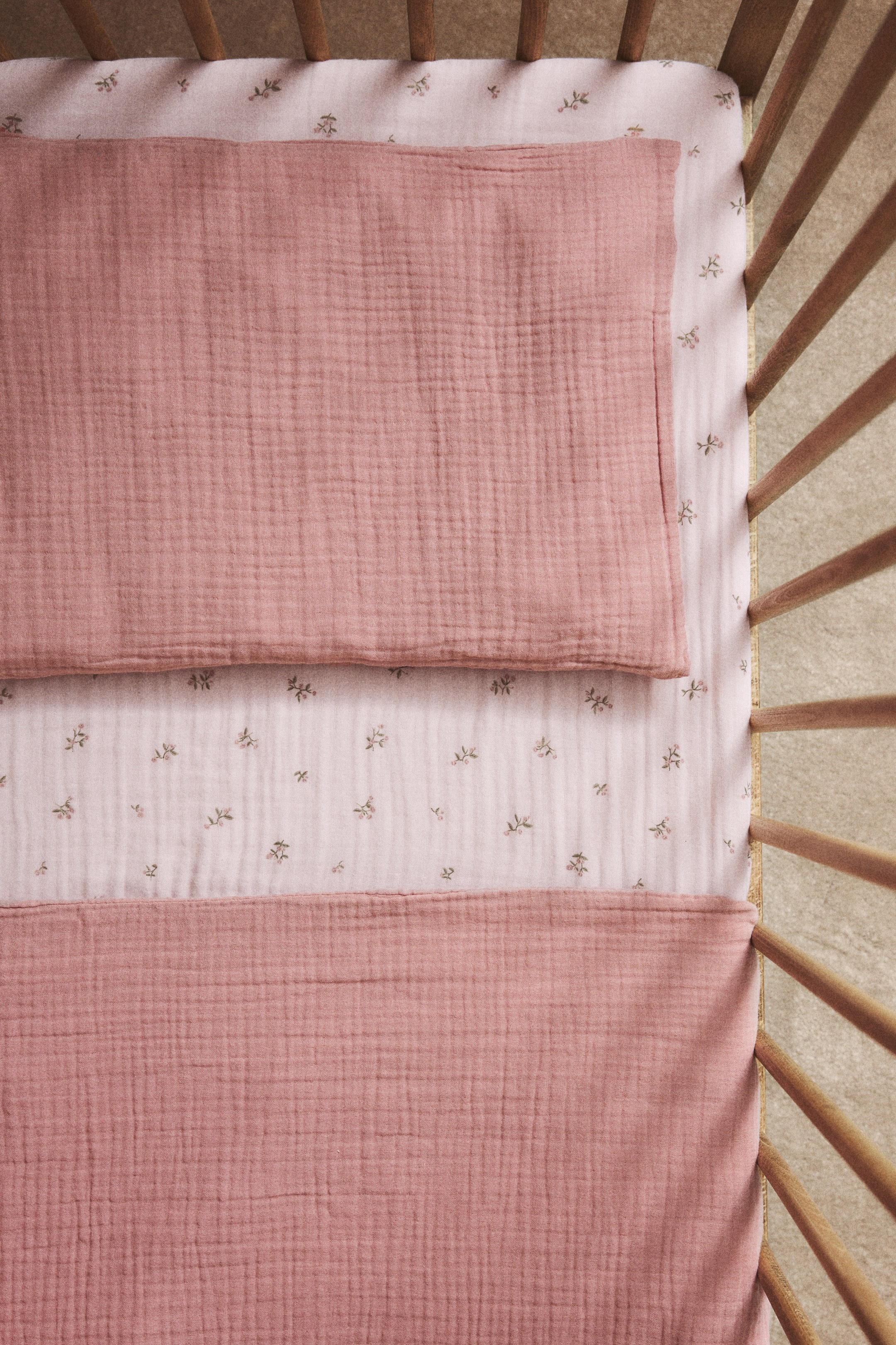 Agrandir l'image: Un drap-housse ajusté en mousseline de coton doux présente un imprimé intégral de petits motifs floraux dispersés sur un fond rose clair. Une couverture en mousseline rose uni et un oreiller assorti reposent sur le drap.