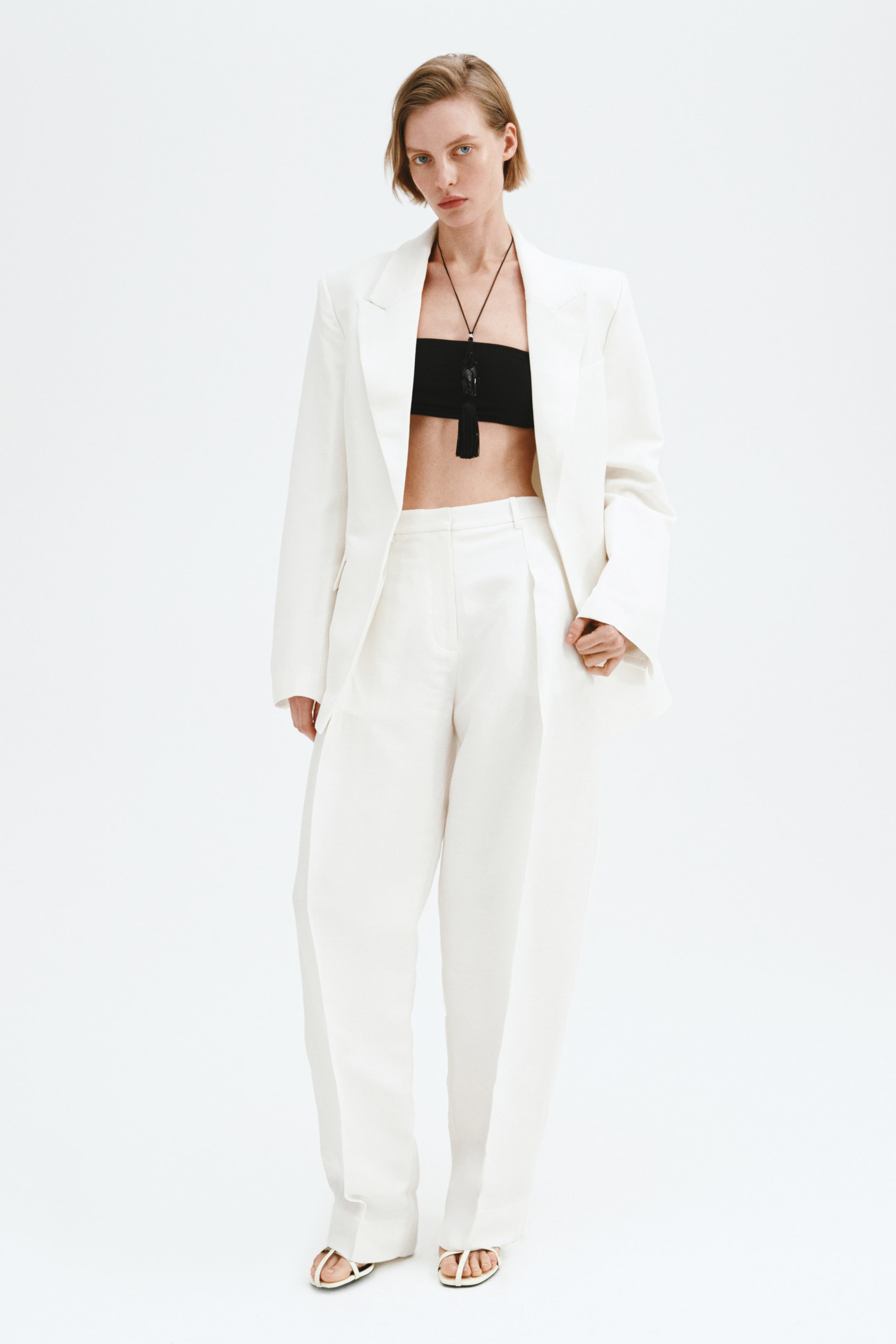 Ingrandisci l'immagine: Un blazer bianco oversize con spalle strutturate e revers a lancia è presentato frontalmente, indossato aperto su un top a fascia nero e una collana con nappine nere. Pantaloni abbinati a gamba larga con pieghe frontali completano l'ensemble, cadendo su sandali infradito bianchi.