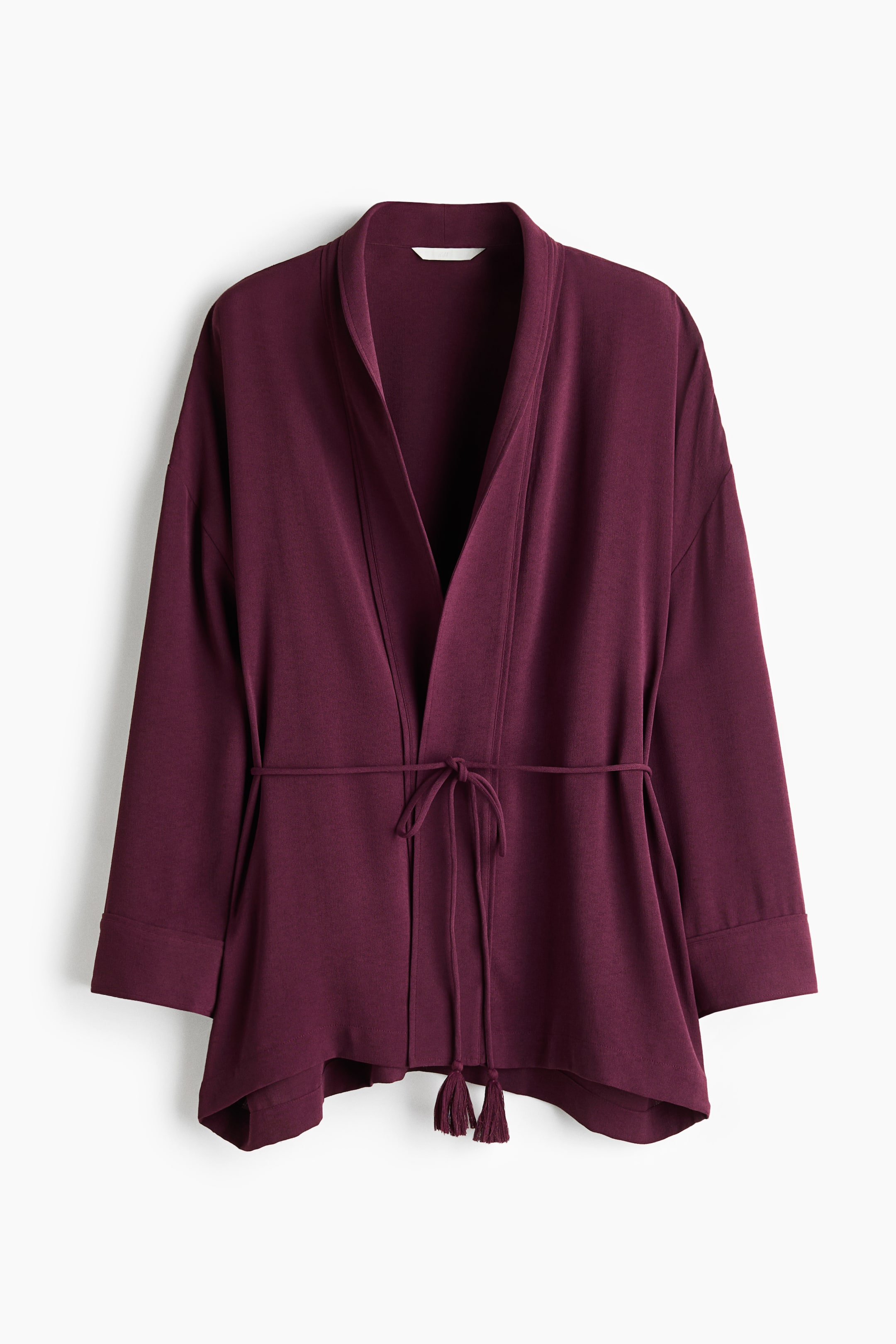 View larger image: A deep plum wrap jacket is hanging front-facing, showcasing an open-front design with a draped shawl collar. The jacket features dropped shoulders, wide-cuffed sleeves, and a thin, tasseled drawstring tie at the waist.