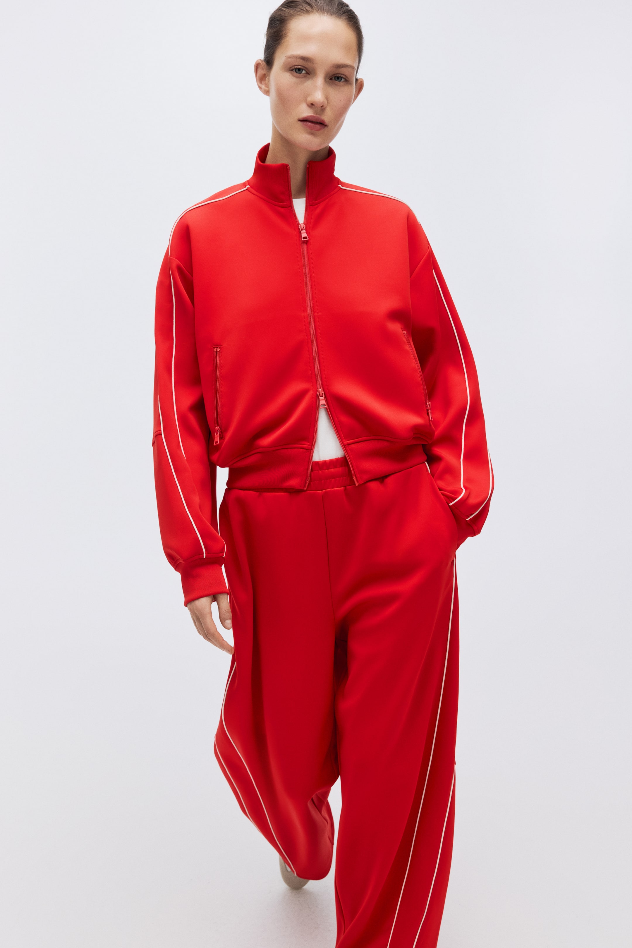 View larger image: A red tracksuit features a zippered jacket with a stand-up collar, ribbed cuffs and hem, and white piping that accents the shoulders, sleeves, and zippered pockets. The jacket is layered over a white T-shirt, and the matching wide-leg pants display white piping along the sides and arcing across the front.