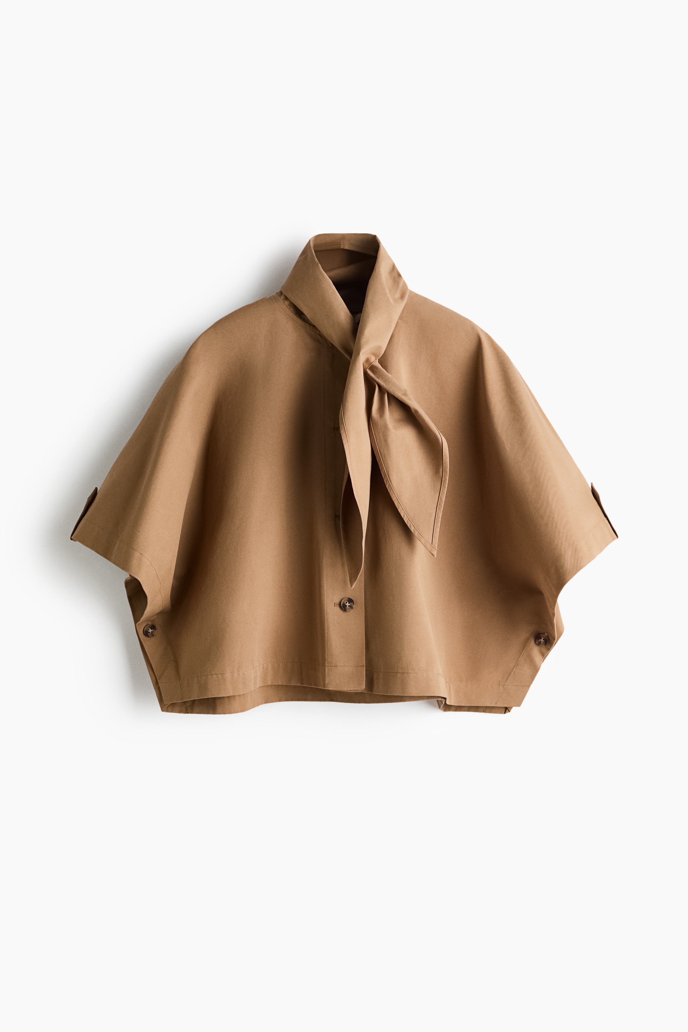 La modelo lleva una capa tipo gabardina de sarga de algodÃ³n beige, con cuello alto tipo bufanda anudado suavemente al cuello. La capa incluye un frente abotonado con botones cafÃ©s visibles y mangas anchas y holgadas.