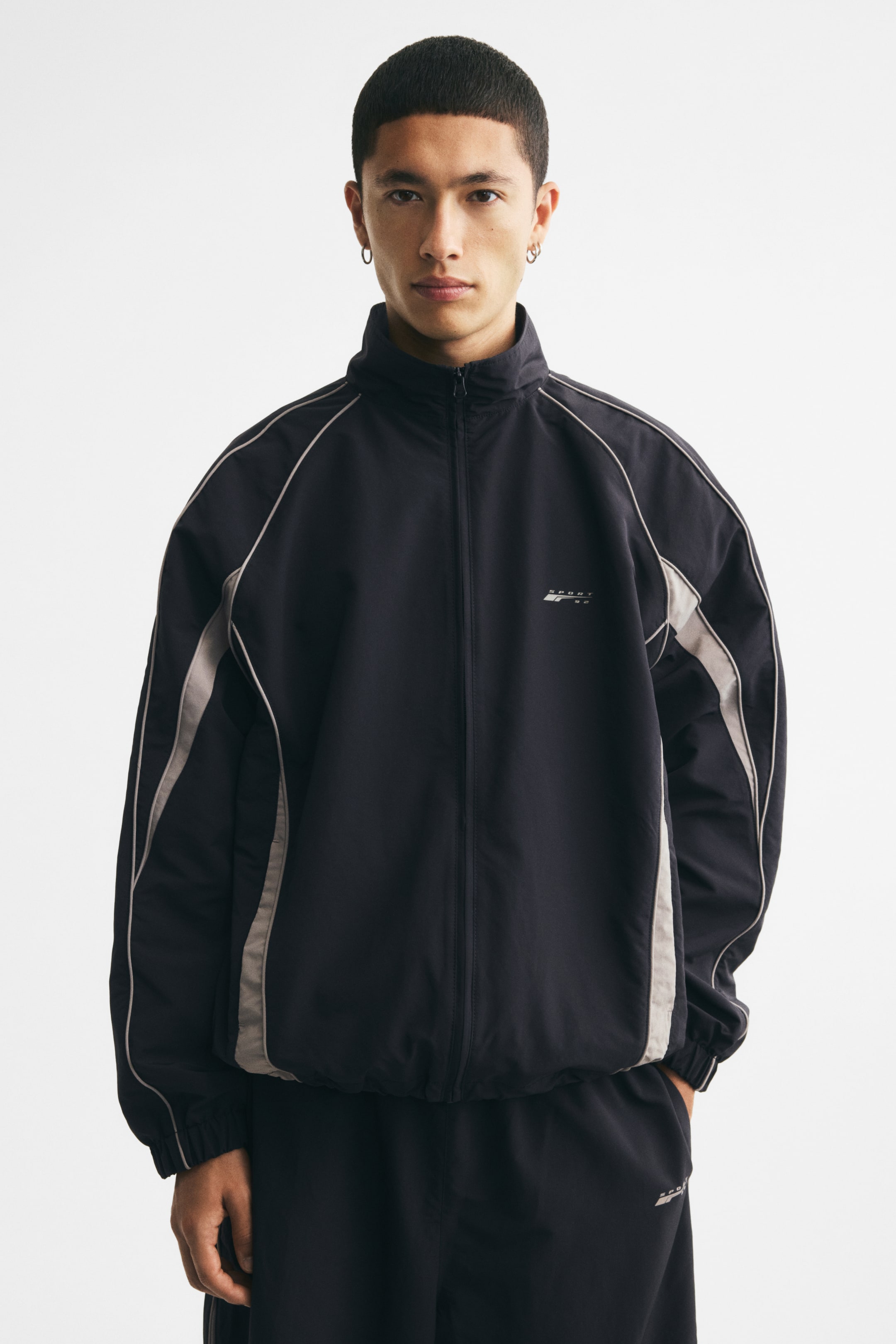 View larger image: The model is wearing a black track jacket with a stand collar, concealed front zip closure, white piping along the shoulders and sleeves, and a logo on the chest. The model’s hands are in the jacket’s side pockets, and the style features covered elastic cuffs and a cinched hem.