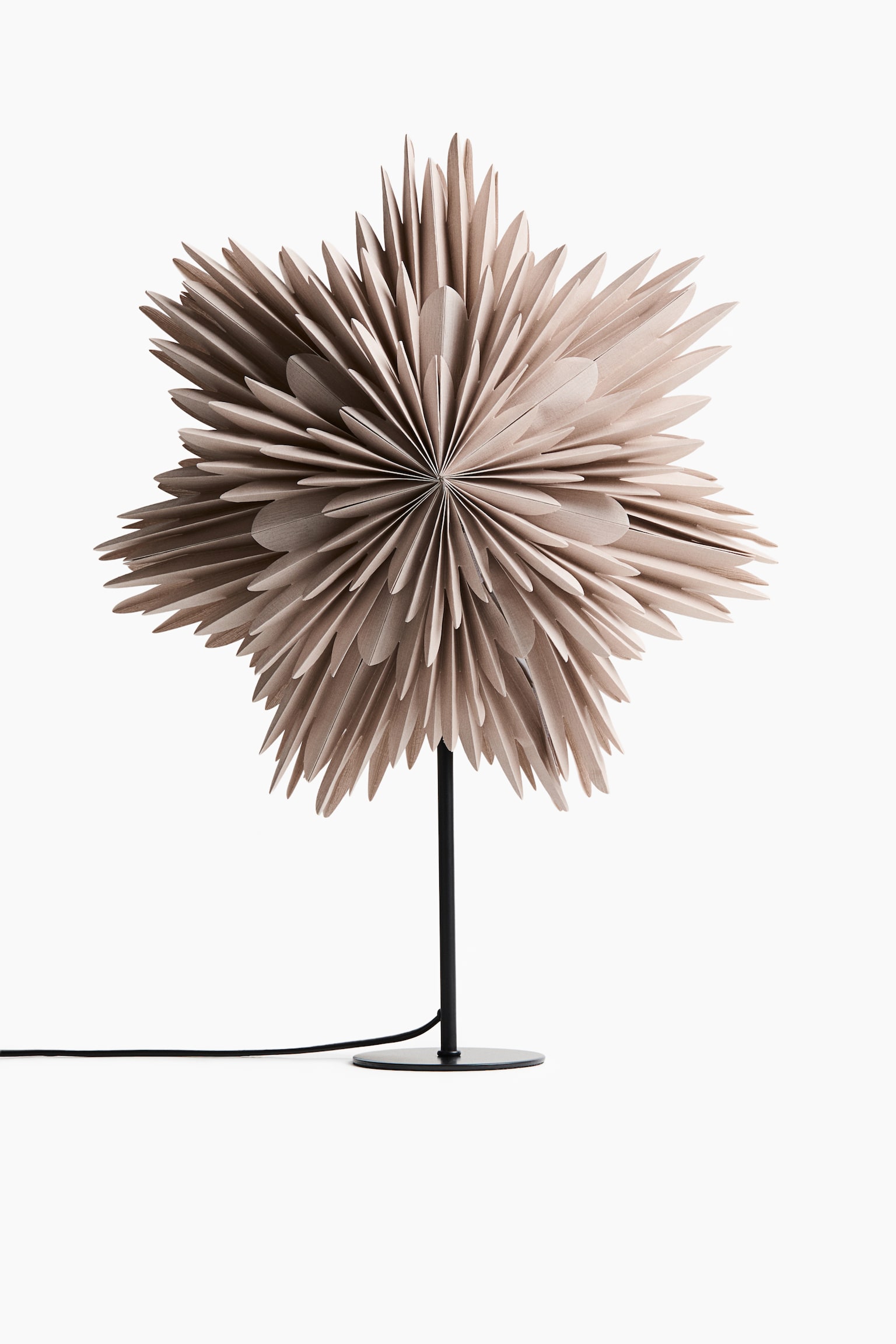 Een tafellamp staat naar voren gericht en heeft een grote, beige papieren kap met dichte, meerpuntige geplooide vouwen die een stervormig patroon vormen. De kap wordt ondersteund door een slanke, verticale zwarte metalen standaard met een platte, ronde voet, met een zwarte voedingskabel die aan de onderkant uitsteekt.