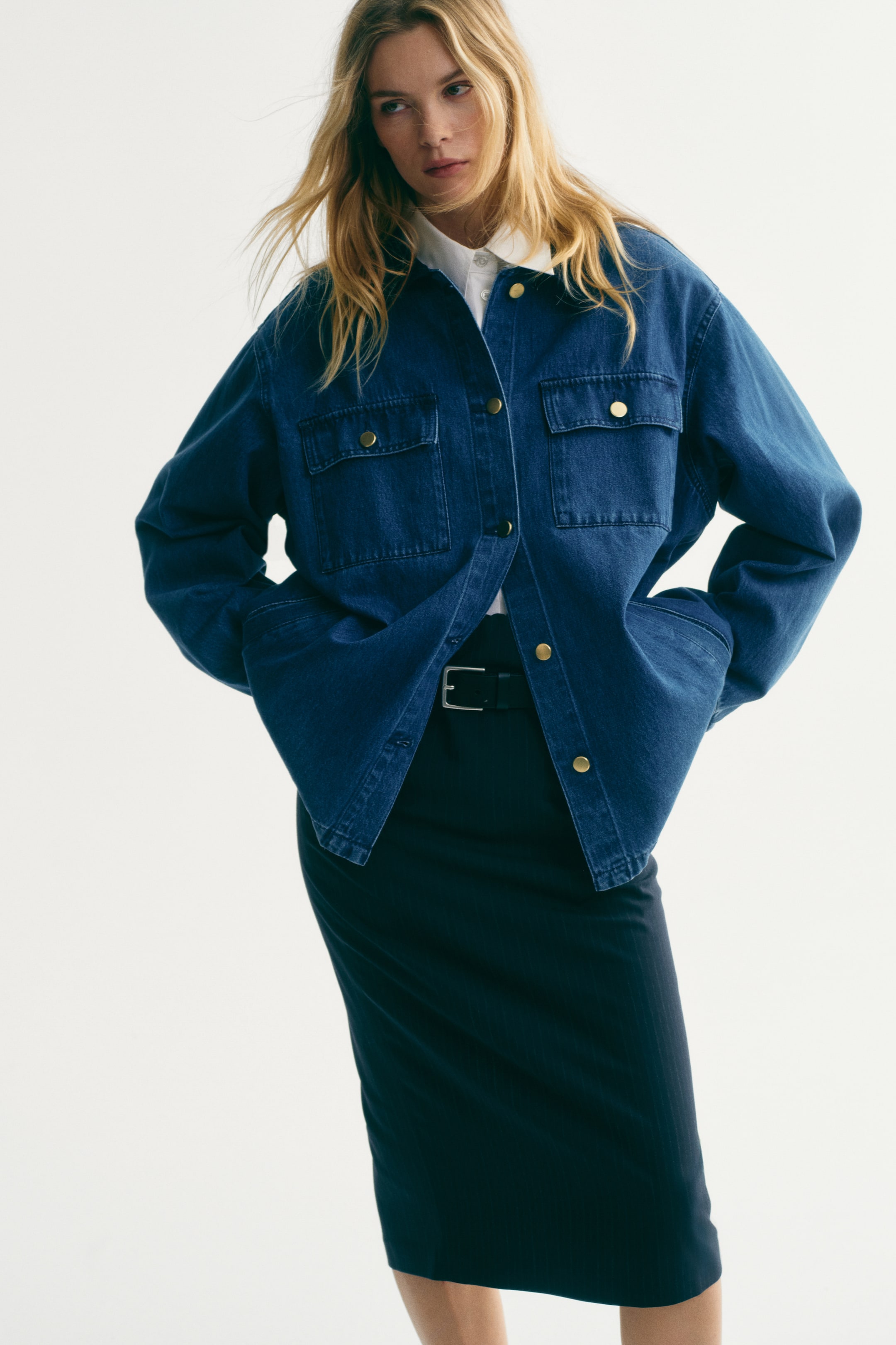 View larger image: A dark blue denim jacket features a relaxed fit, with gold-toned buttons down the front and two chest flap pockets. It is styled open over a white collared shirt, with hands resting in its side pockets.