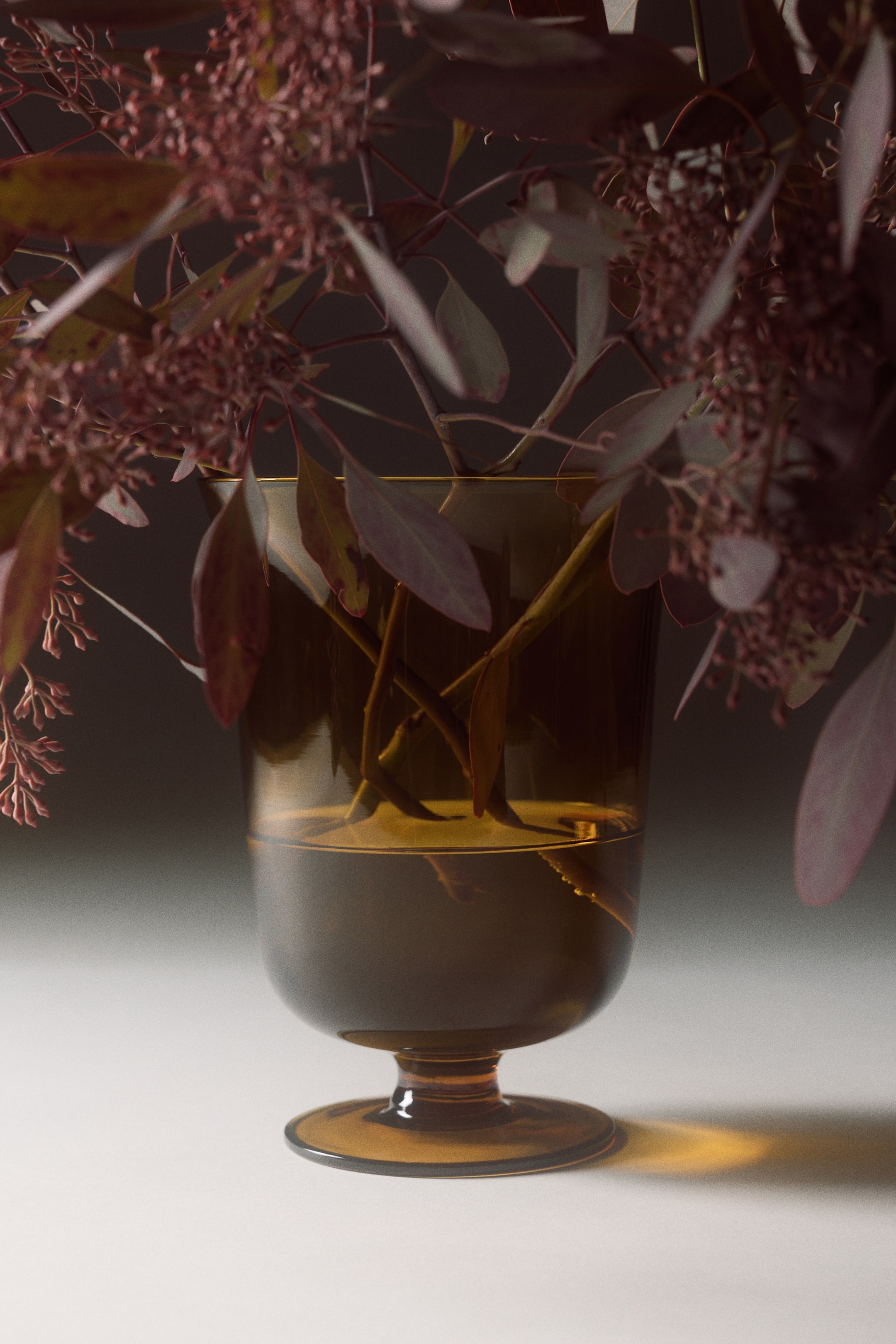 View larger image: A glass footed vase made of translucent amber glass is shown from the front, featuring a gently flared opening, a rounded body, and a short stem on a flat circular base. Several leafy branches with reddish-purple berries are resting inside the water-filled vase, highlighting its transparency and timeless design.
