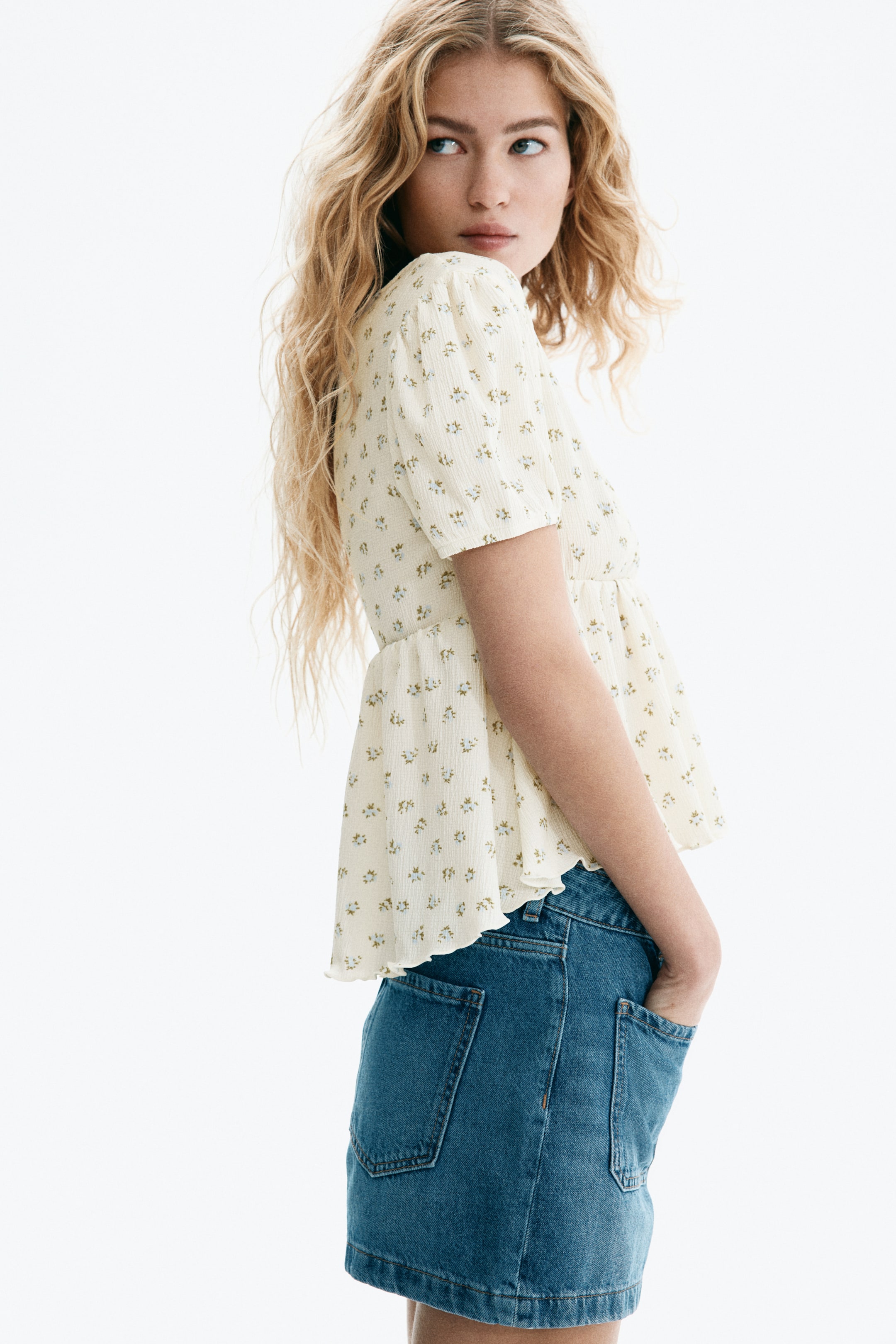View larger image: A cream-coloured peplum top features a subtle textured fabric with a delicate floral print, short puffed sleeves and a gathered waist that flares at the hem. It is styled with a mid-blue denim mini skirt with visible back pockets and a straight silhouette.