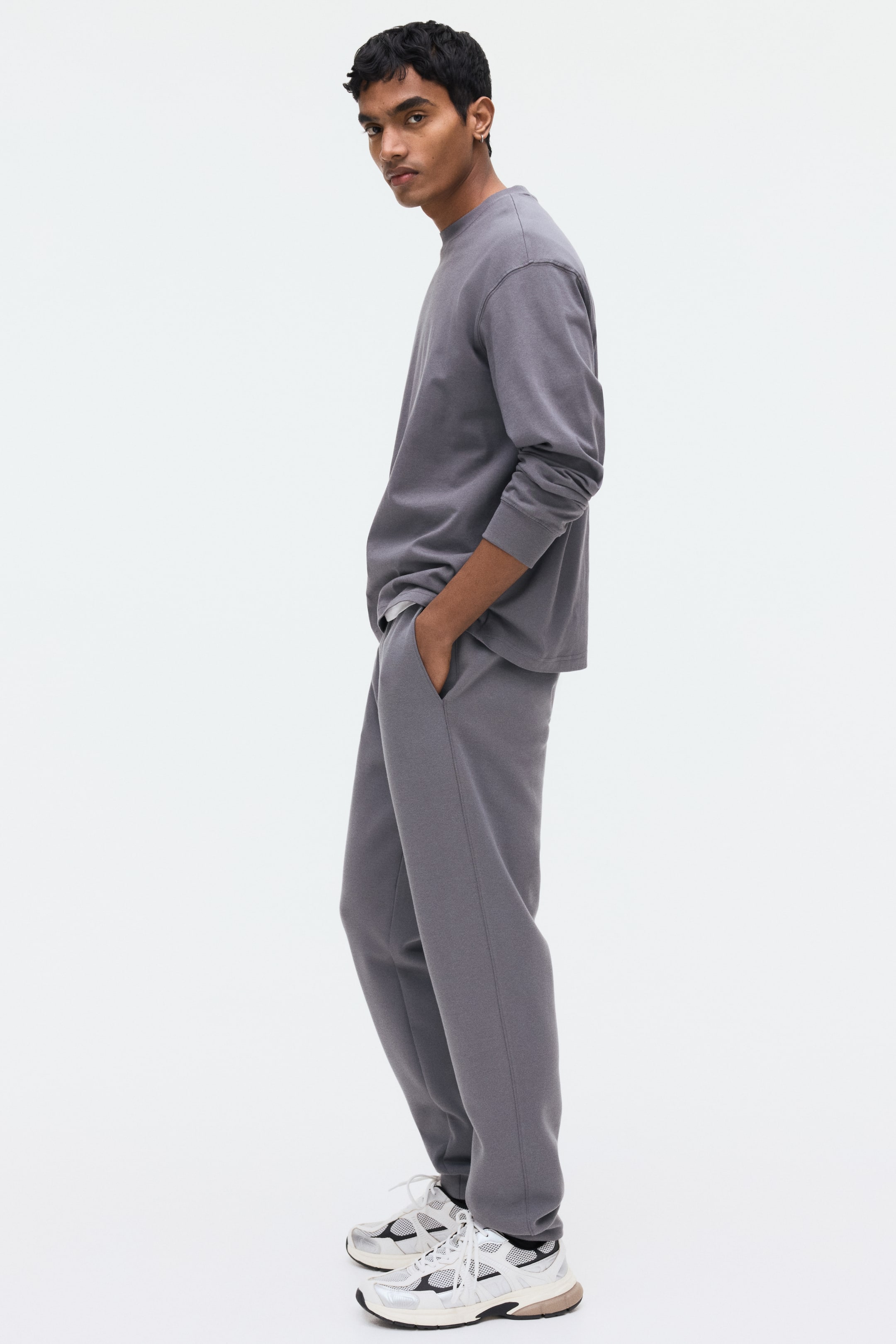 Ingrandisci l'immagine: I pantaloni della tuta grigi presentano una vita elasticizzata, tasche laterali diagonali e gambe affusolate con costine agli orli. Il modello indossa i pantaloni della tuta, abbinati a un top a maniche lunghe coordinato, e mette una mano in una tasca laterale.