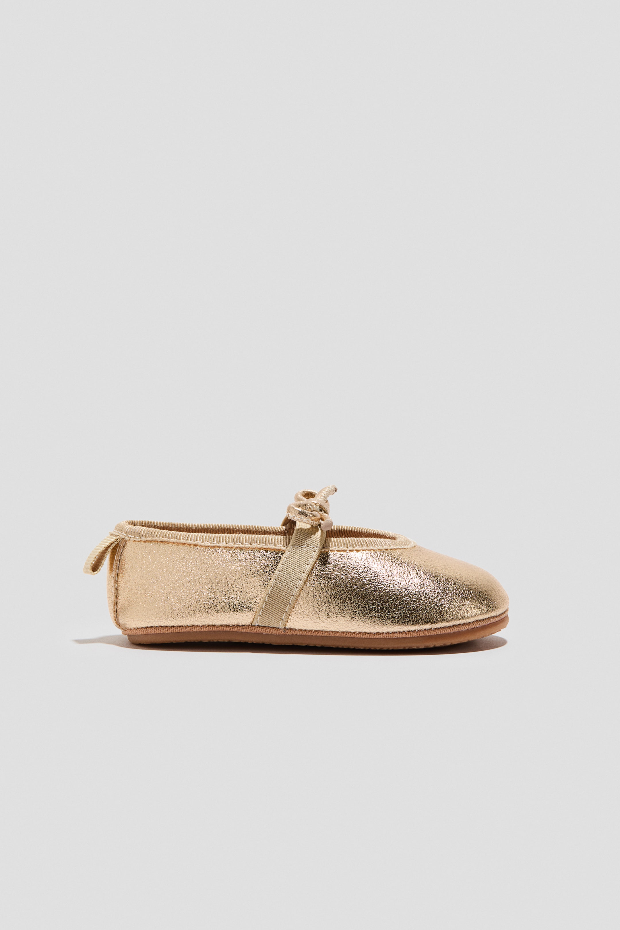 View larger image: A gold ballet pump showcases a rounded toe and a grosgrain strap elegantly crossing over the foot, tied into a neat little knot. It also features a grosgrain loop at the back and a flat, brown sole.