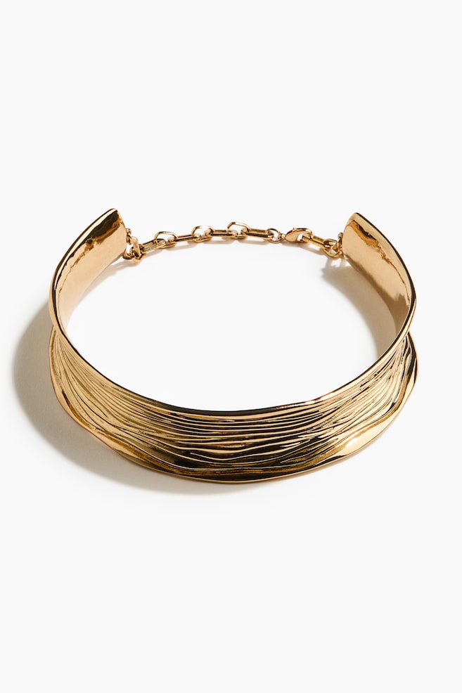 A gold-toned collar necklace is presenting a wide, layered design on its front, composed of multiple wavy, thin bands. It is featuring a link chain extender and a clasp at its ends. - 1