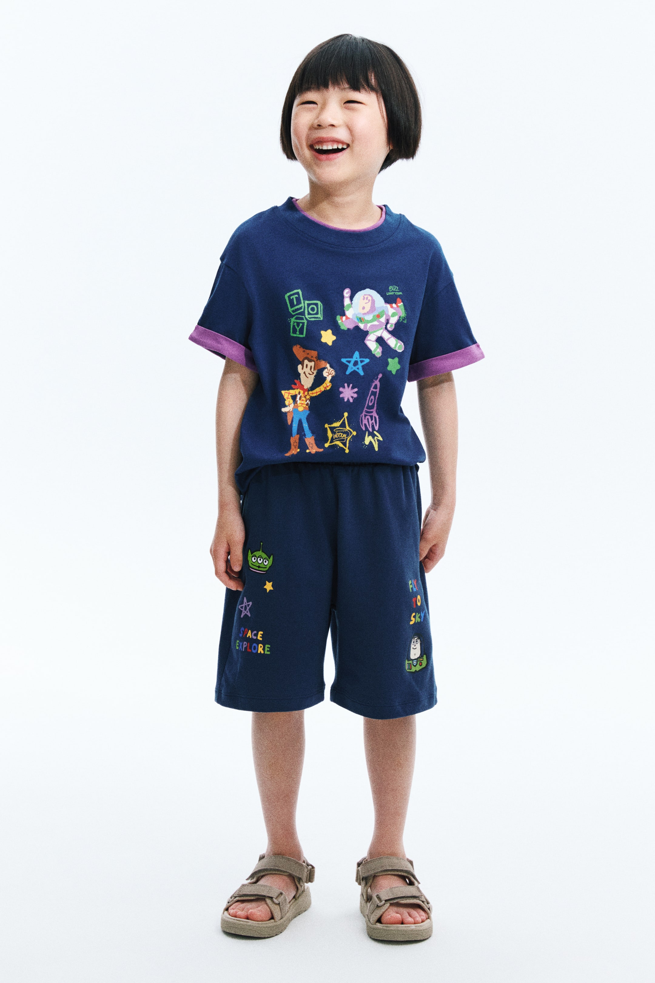 View larger image: A navy blue children's two-piece outfit is being presented, facing forward. The short-sleeved t-shirt is displaying Woody, Buzz Lightyear, "TOY" text, a rocket, and stars, with purple cuffs and a visible purple layer at the crew neck. The knee-length shorts are displaying an alien, "SPACE EXPLORE" text, and Forky with "FLY TO SKY" text.