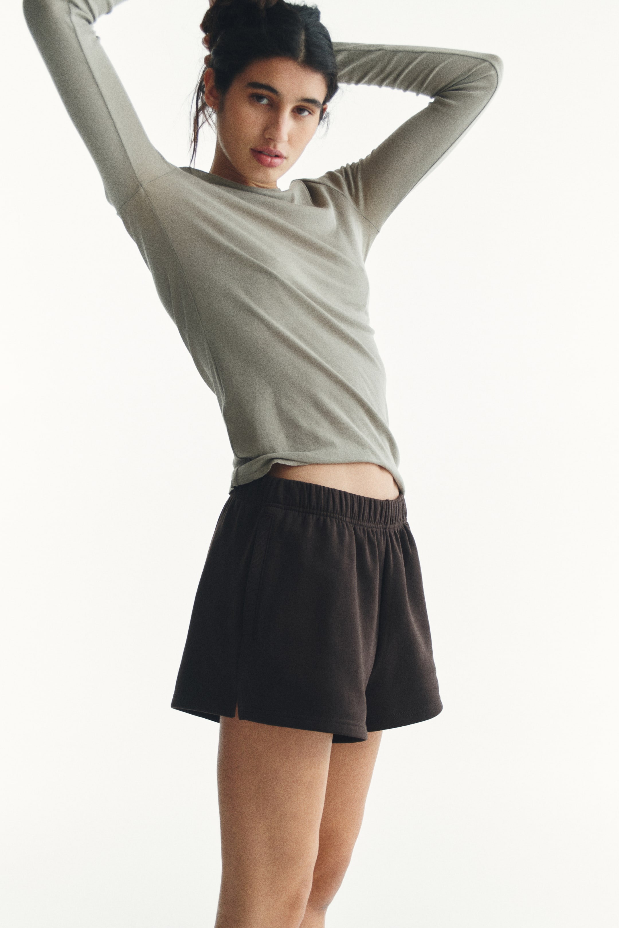 View larger image: A light gray, fitted, long-sleeve top with a round neckline and sleeves extending overhead is paired with dark brown shorts, featuring an elastic waistband, side pockets, and short side slits.