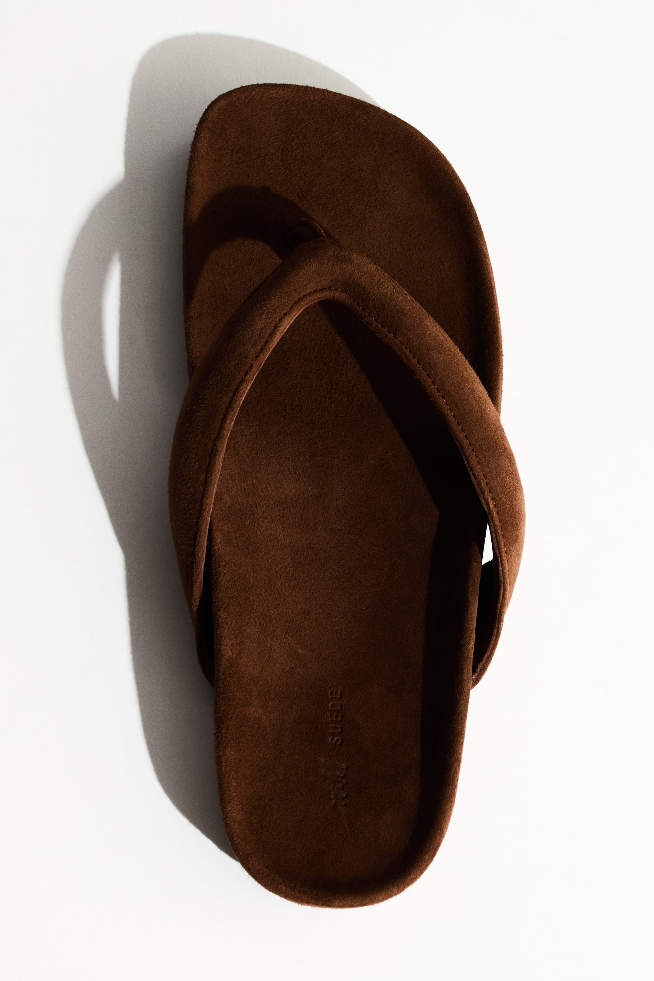 View larger image: A brown suede flip-flop is displayed from a top-down perspective, highlighting an oblique toe, cushioned straps, and a slender toe post.