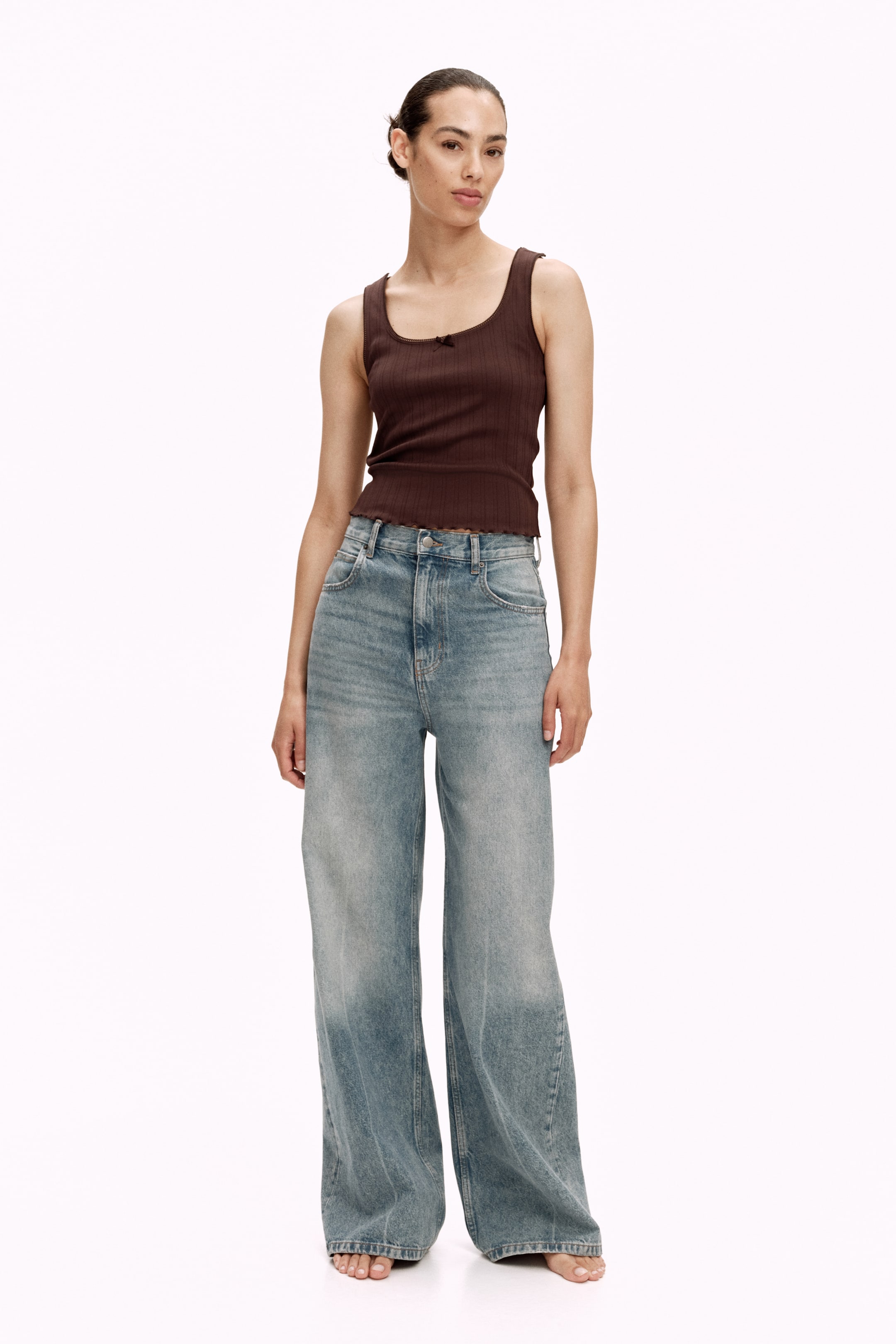Größeres Bild ansehen: Ein dunkelbraunes, geripptes Tanktop wird von vorne präsentiert und verfügt über einen Rundhalsausschnitt mit einer kleinen Schleifendetail. Es zeigt eine figurbetonte Silhouette und einen gewellten Saum, der knapp über hell gewaschenen Jeans liegt.