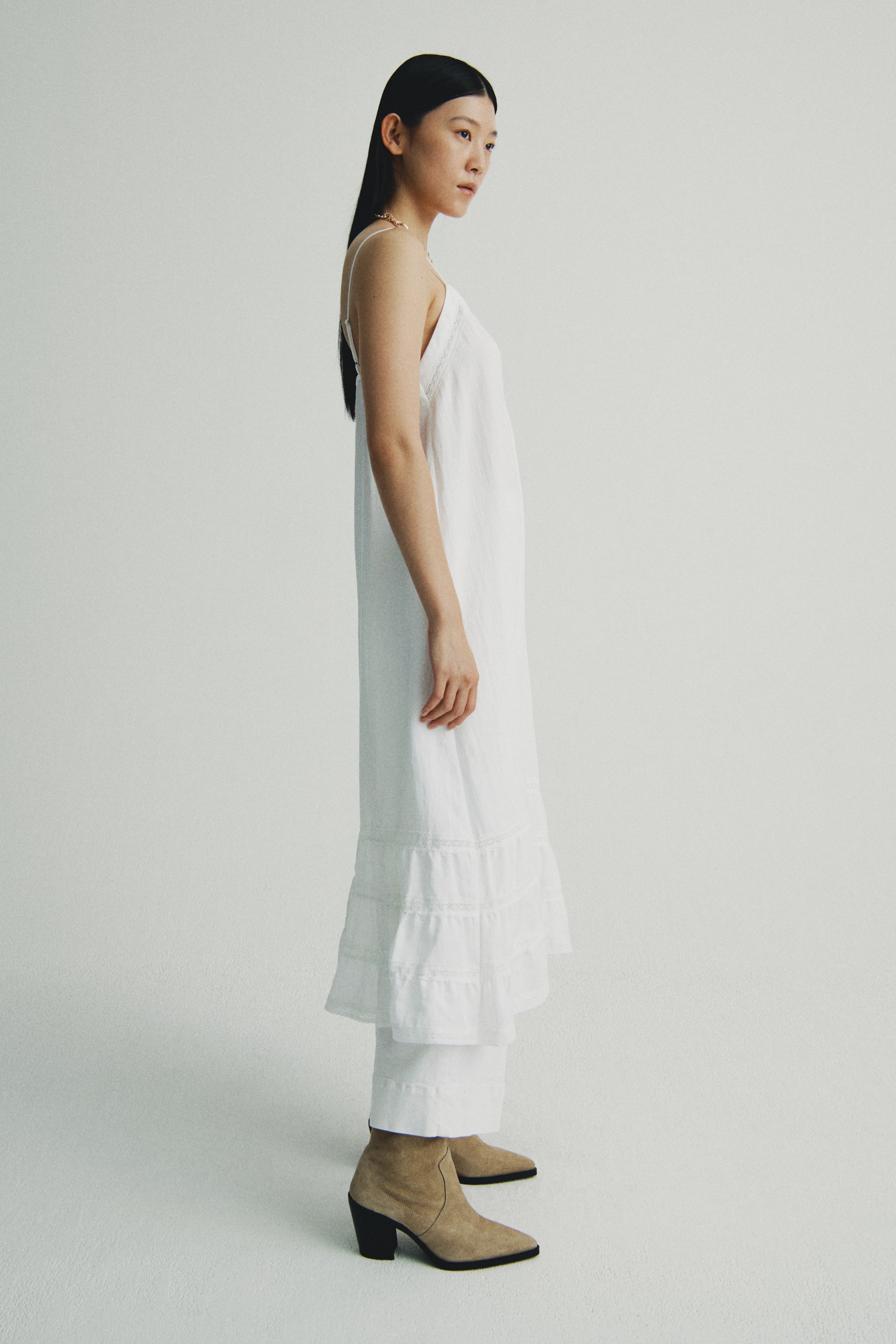 Xem ảnh lớn hơn: Một chiếc váy maxi trắng không tay với dây đeo mảnh và gấu váy xếp tầng có viền ren, được mặc bên trên quần trắng dáng suông, nhìn từ góc nghiêng. Đôi bốt cổ thấp màu nâu da lộn với mũi nhọn và gót vuông có thể nhìn thấy trên chân.