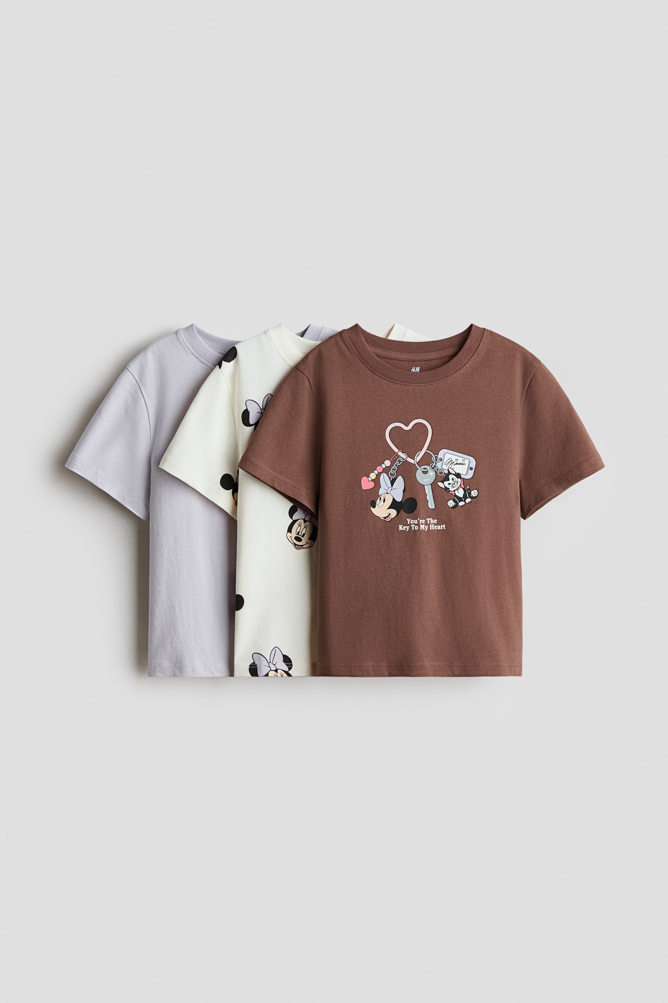 View larger image: A brown short-sleeved t-shirt is showcased from the front, adorned with a graphic of Minnie Mouse, a dog, and keys on a heart-shaped keychain, accompanied by the text "You're The Key To My Heart." In the background, a cream t-shirt flaunts an all-over black Minnie Mouse head print, while a light purple t-shirt is simply plain.