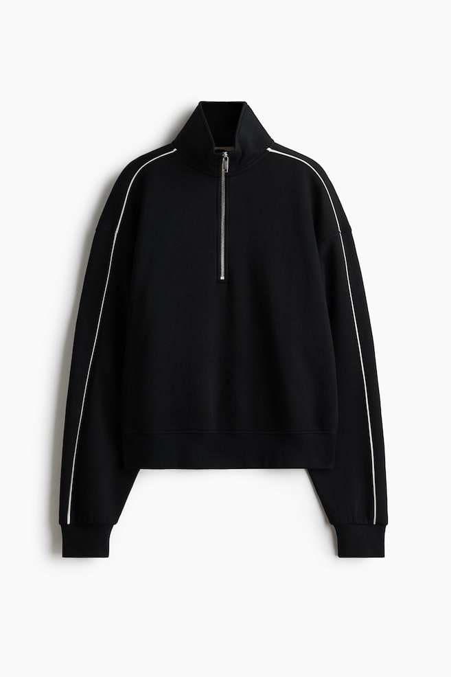 En sort sweater med halv lynlÃ¥s ses forfra og har en stÃ¥ende krave med en sÃ¸lvfarvet metallynlÃ¥s. Tynde hvide kantbÃ¥nd lÃ¸ber ned langs de sÃ¦nkede skuldrer og de lange Ã¦rmer, som afsluttes med ribkanter over en ribbet kant forneden. - 1