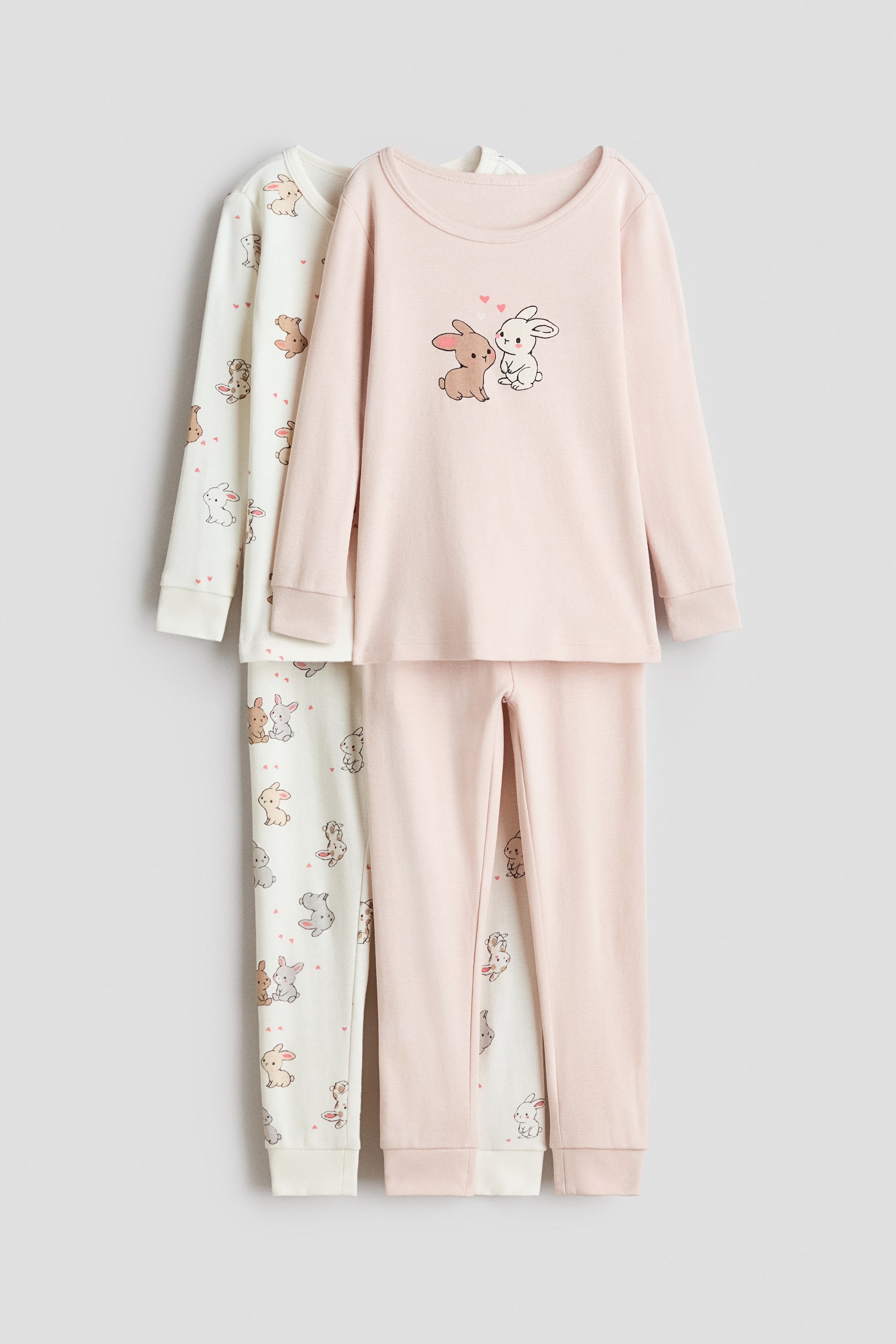 View larger image: Two sets of pajamas are being presented, both including long-sleeved tops with ribbed necklines and cuffs, and bottoms with elasticated waists and ribbed hems. The front pink set is displaying a graphic of two rabbits with hearts on its top, while the cream set positioned behind it is patterned with multiple small rabbits and hearts.